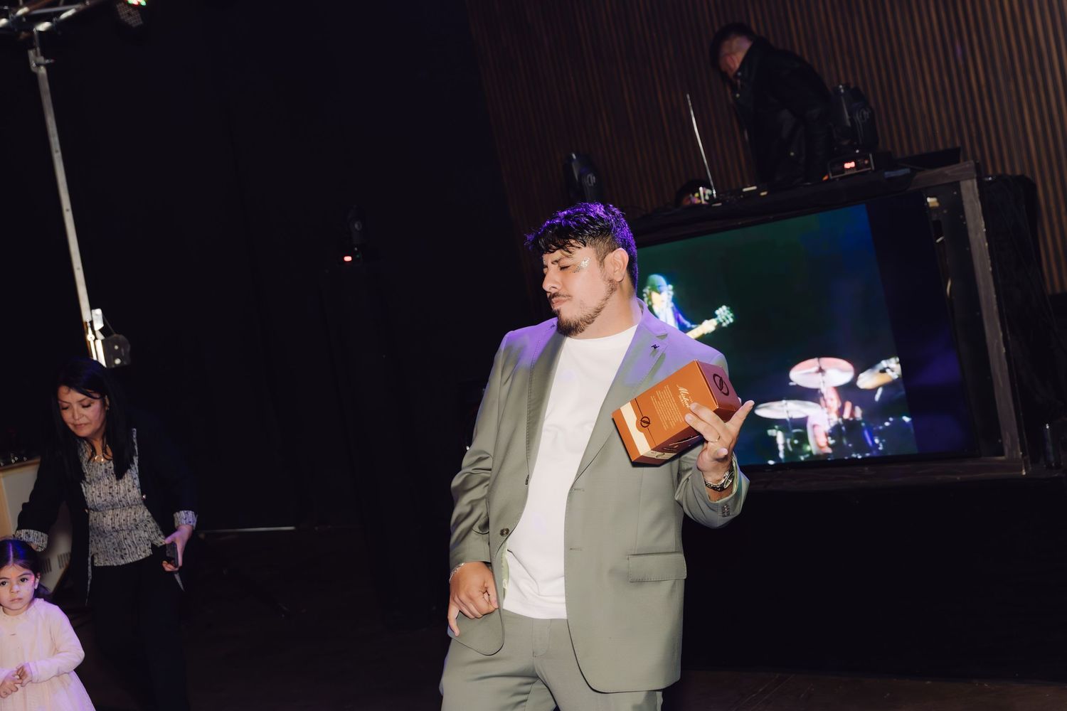 Young man in gray suit holding wooden gift box near stage at wedding reception.