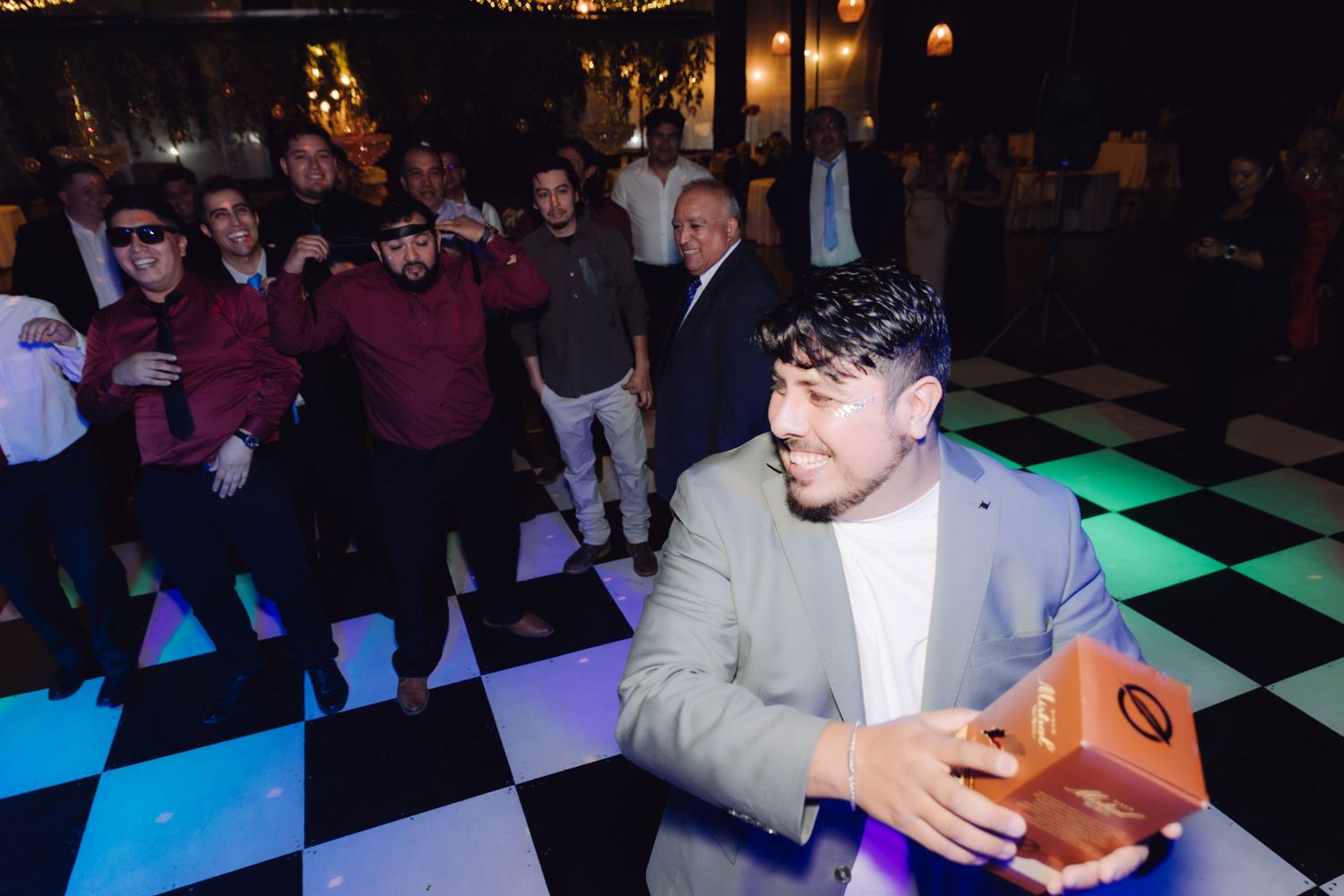 Man in gray blazer holding orange gift box on checkered dance floor at wedding.