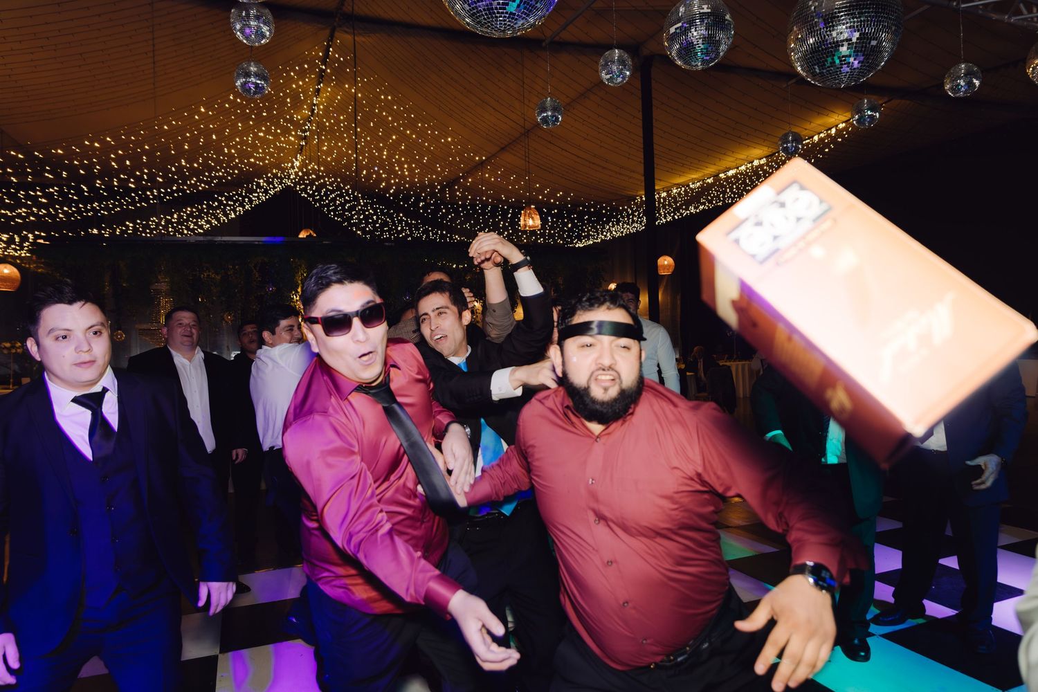 Guests posing and dancing with gift box on checkered dance floor at wedding party.