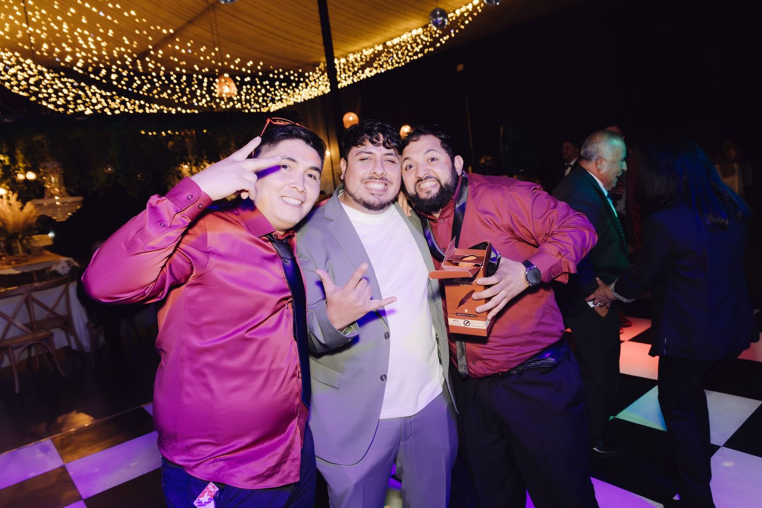 Three men posing together on dance floor at lively wedding reception under string lights.