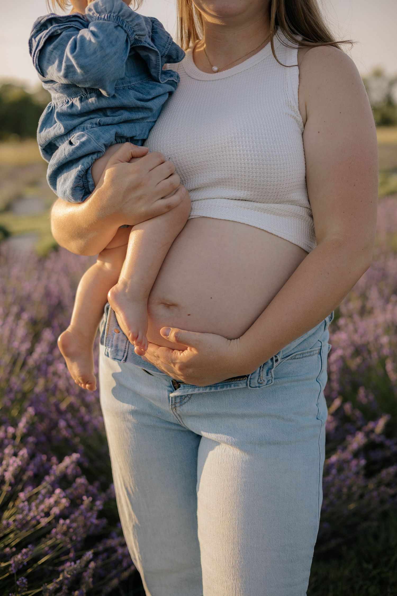 Pregnant woman in white crop top and jeans holds a baby in a lavender field at sunset.
