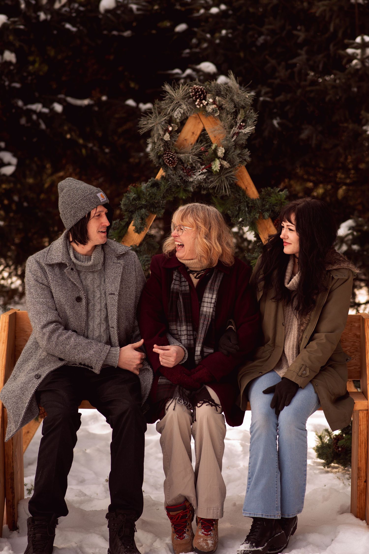 Three people sitting on a bench in the snow, laughing together in front of a Christmas wreath outdoors.