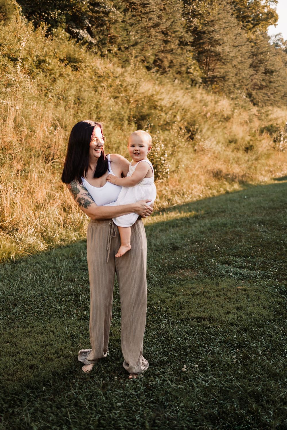 A smiling mother with dark hair and tattoos holds her happy baby outdoors in golden hour light on a green lawn in Knoxville, TN.