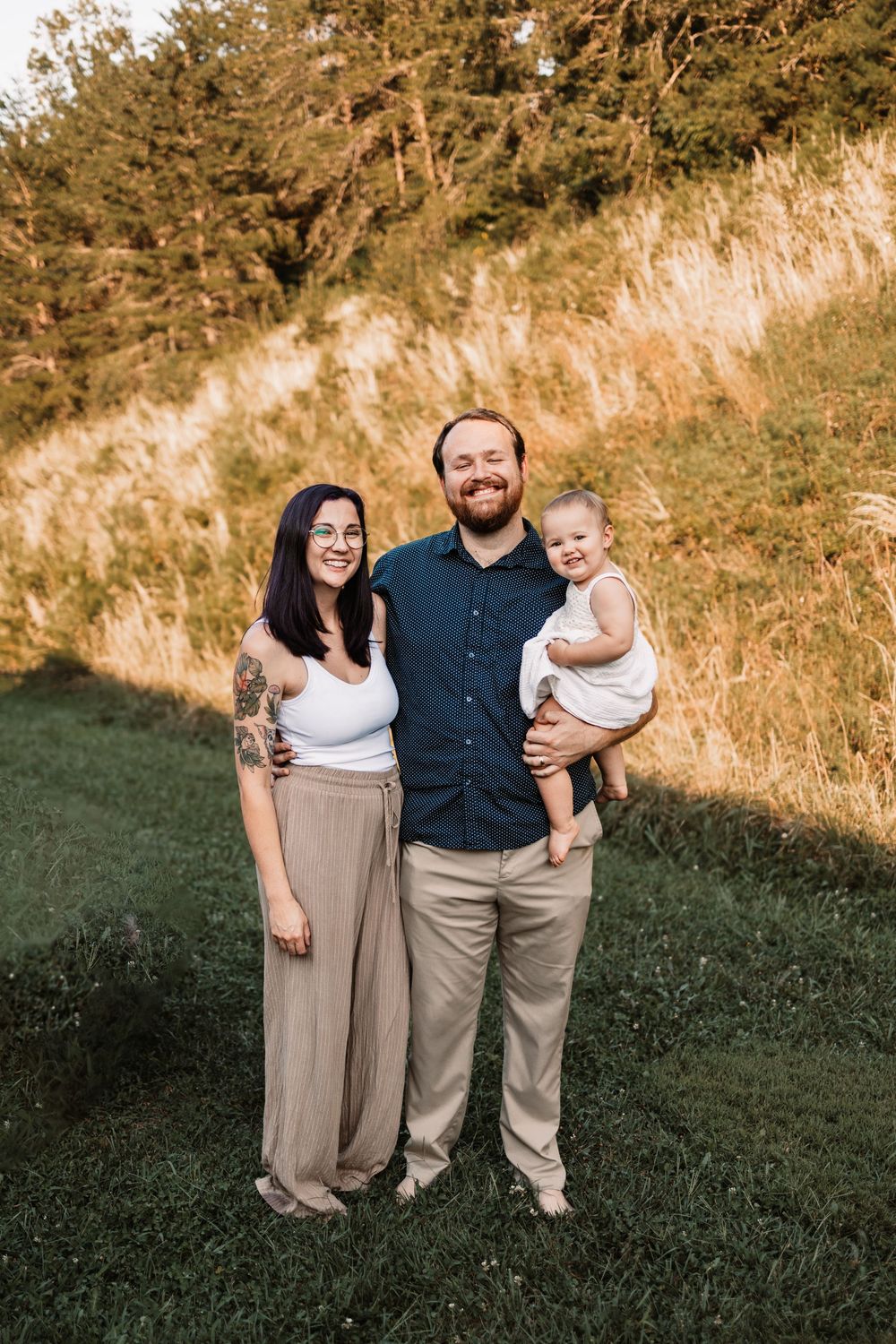 Happy family portrait outdoors at golden hour, parents smiling with baby in white outfit on green grass in Knoxville, TN.