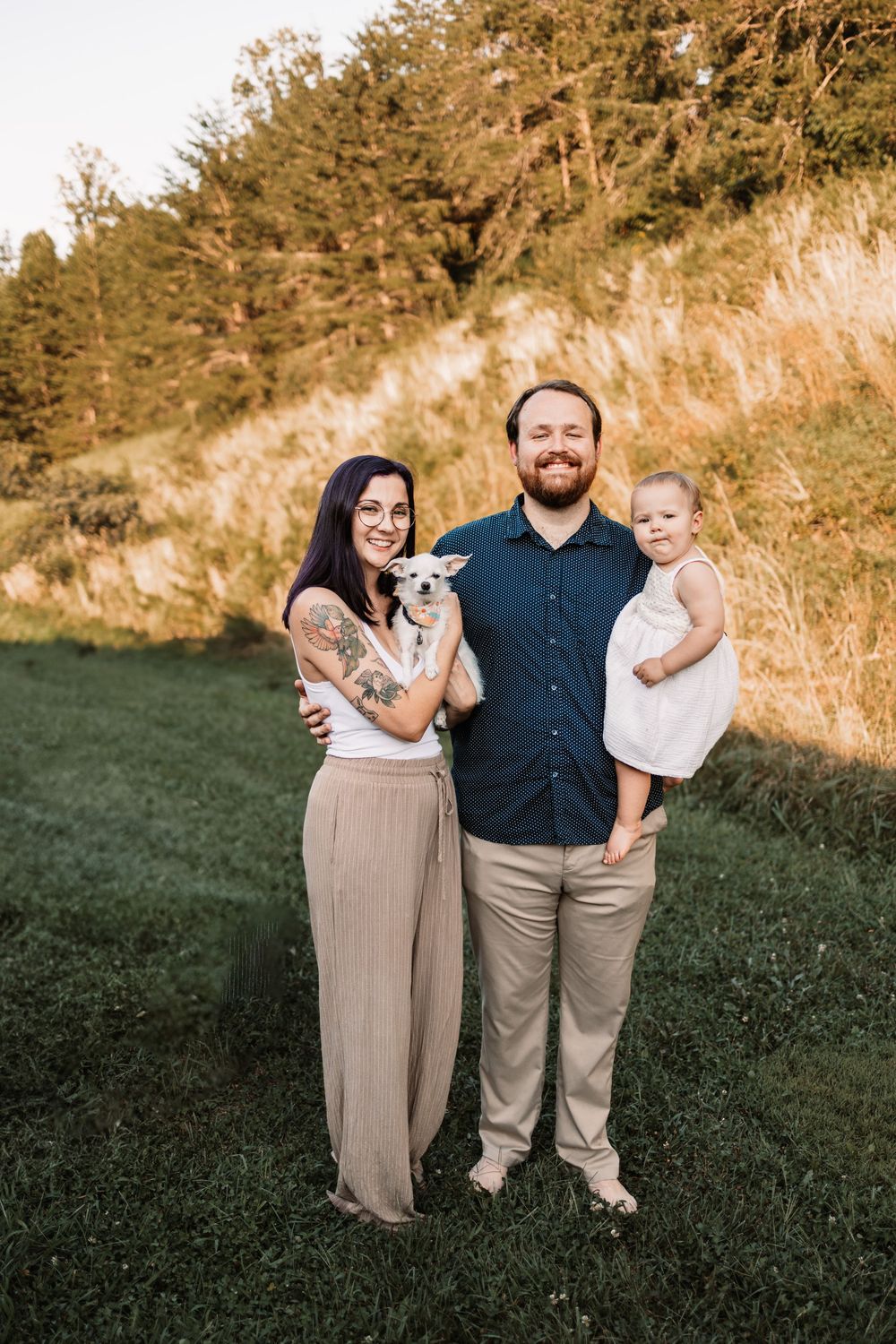 A smiling family of three poses outdoors, with a woman holding a small dog and a man carrying a toddler girl in Knoxville, TN.