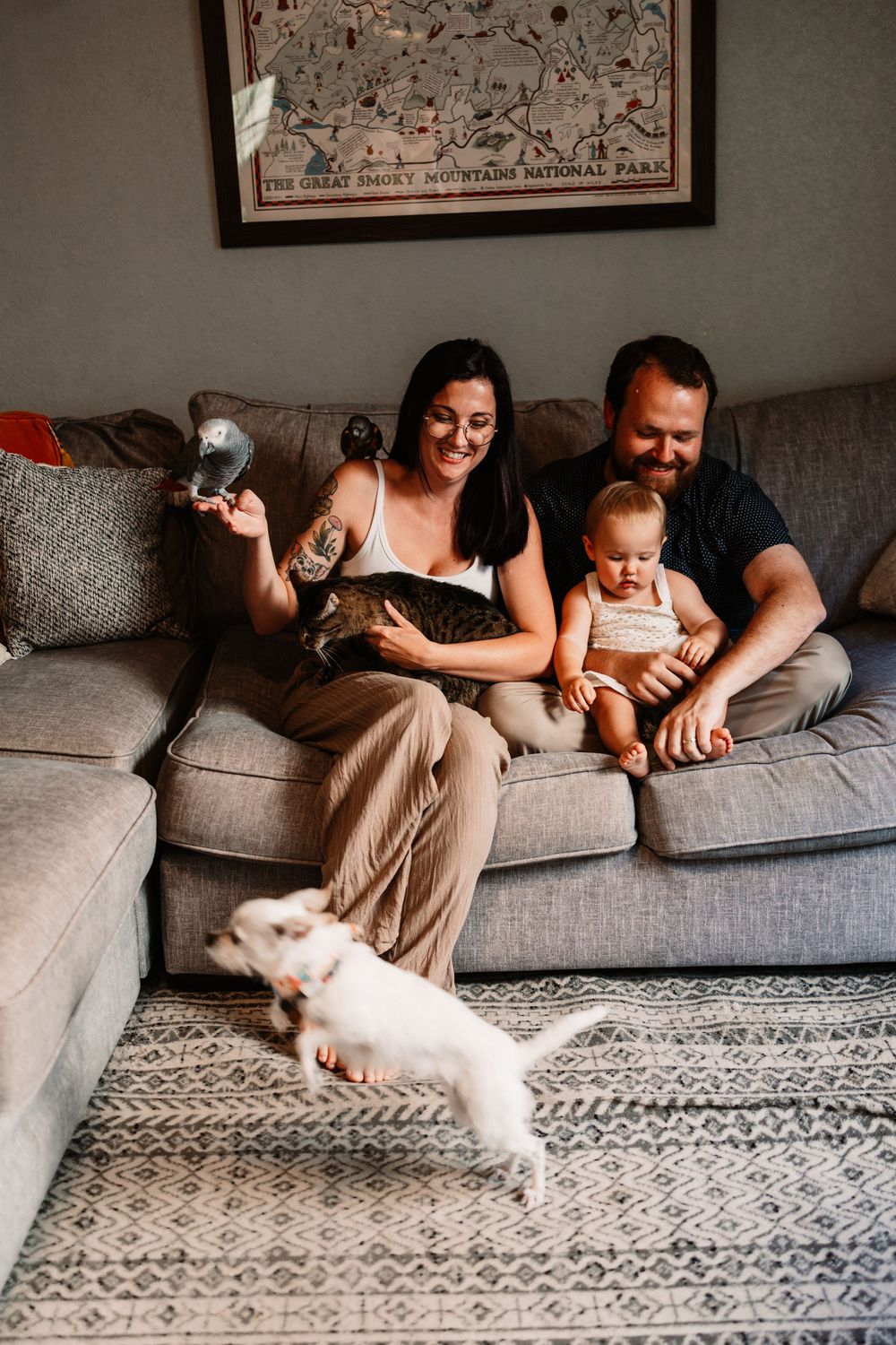 Family with baby and cat sit on gray sofa while white dog plays on patterned rug below Great Smoky Mountains map in Knoxville, TN.