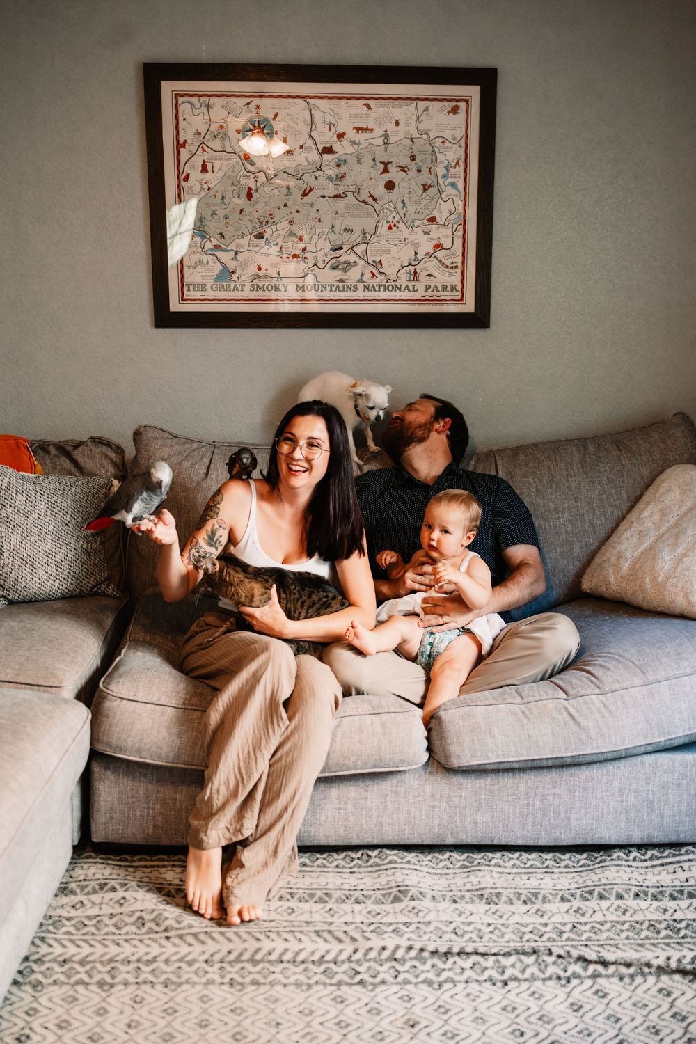 Happy family with baby and parrot relaxing on gray sectional sofa beneath Great Smoky Mountains National Park map in Knoxville, TN.