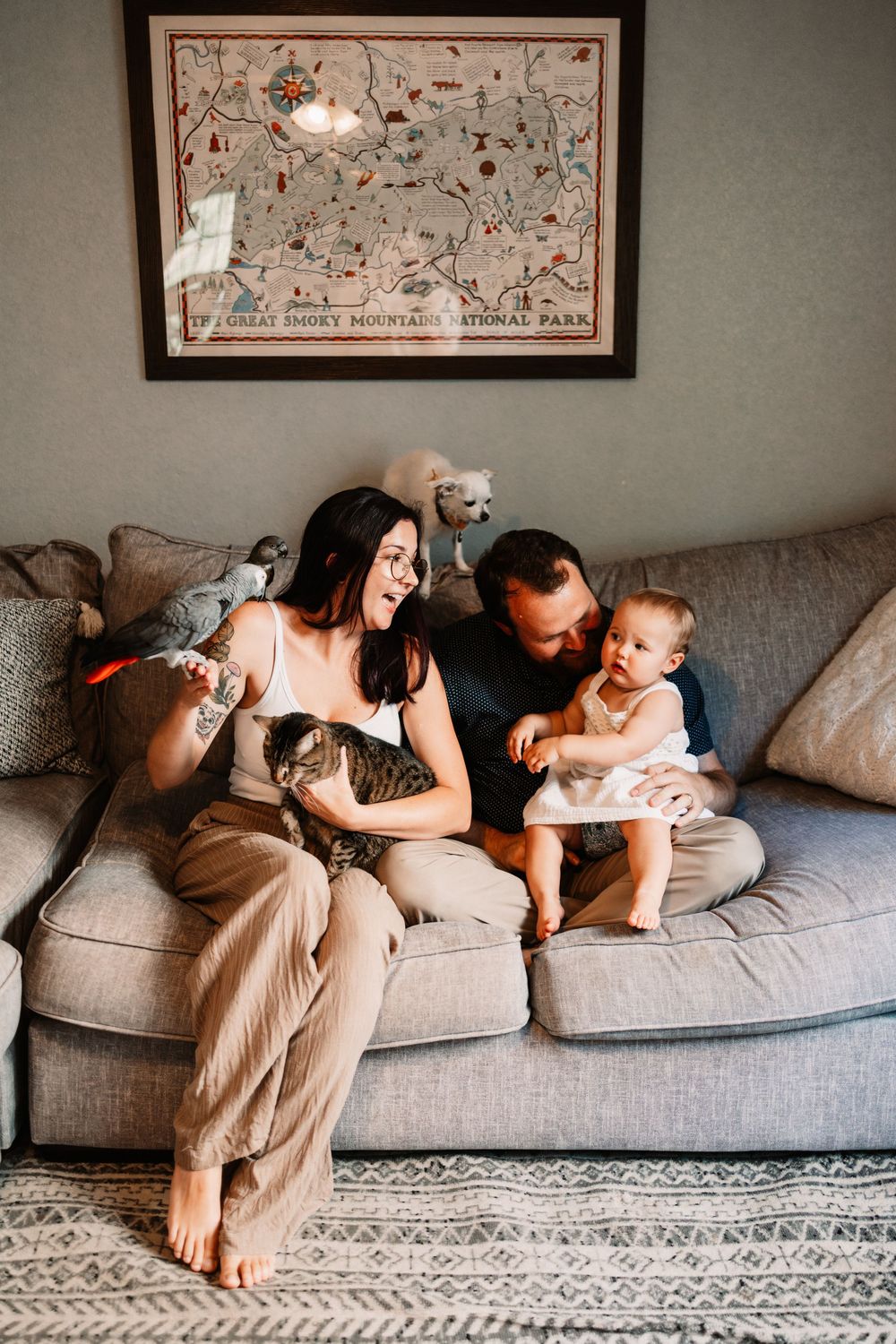 A young family sits on a gray couch with a tabby cat and baby, beneath a Great Smoky Mountains National Park map in Knoxville, TN.