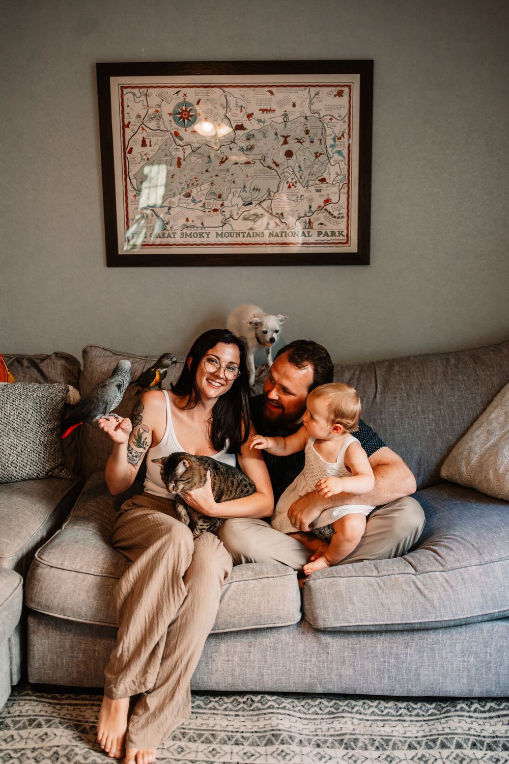 A happy family with a bearded man, smiling woman with tattoos, baby, and cat sit on a gray couch beneath a framed map in Knoxville, TN.