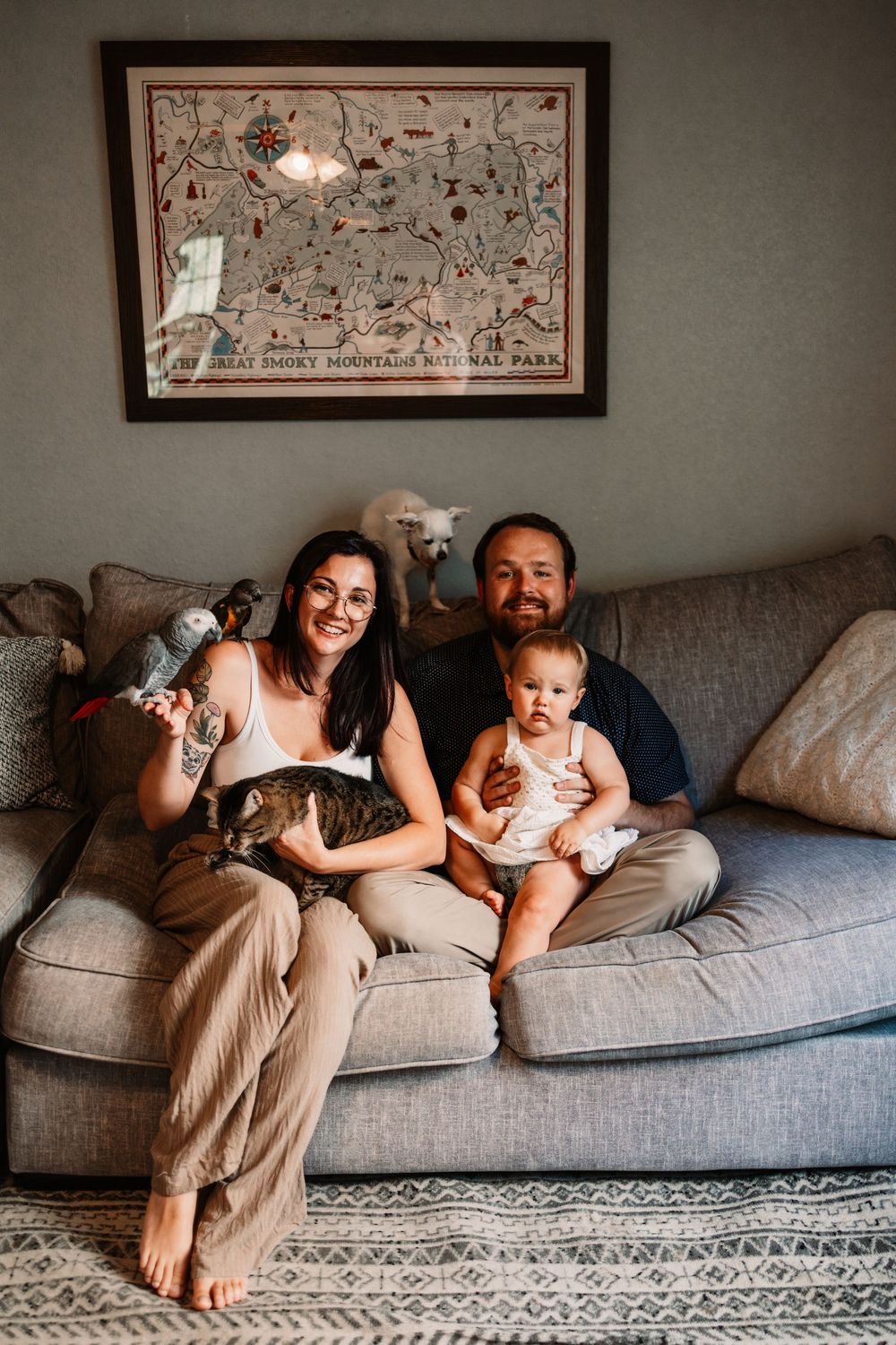Family of three with a baby, cat, and parrot sitting on a gray couch beneath a Great Smoky Mountains map in Knoxville, TN.