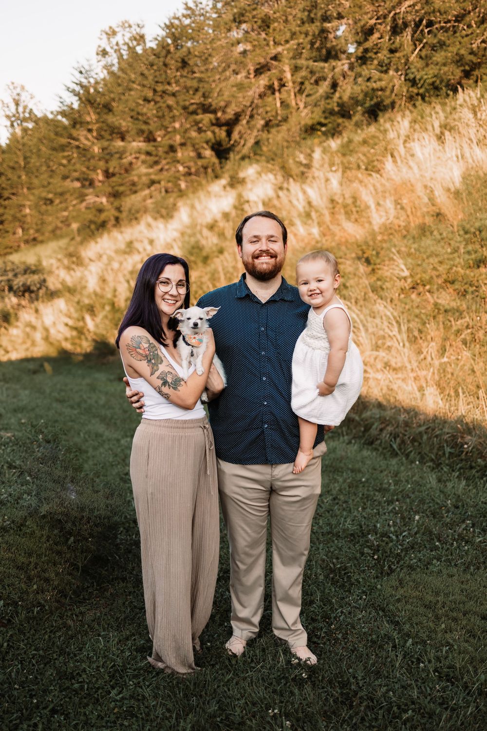 Family portrait of couple with small white dog and toddler girl on green hillside at golden hour in Knoxville, TN.