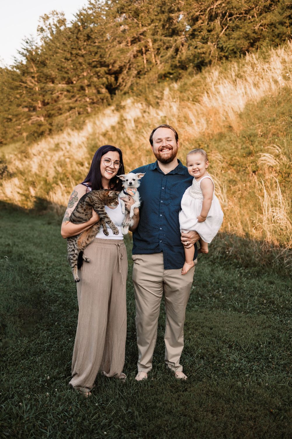 A smiling family poses outdoors holding a cat and small dog, with a toddler in white dress, set against a golden hillside in Knoxville, TN.