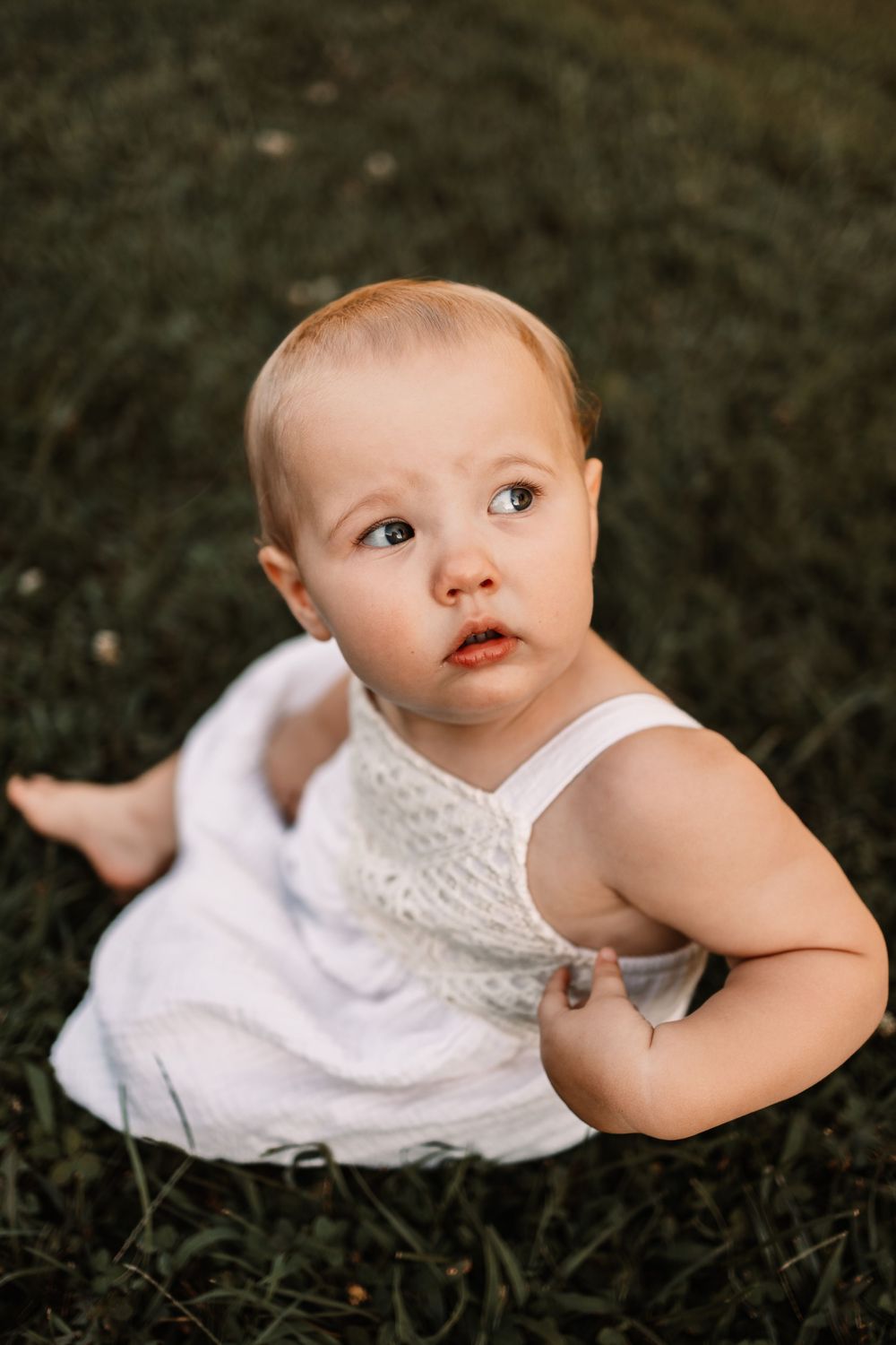 A cute baby in a white dress sits on green grass, looking up with big brown eyes in an outdoor portrait in Knoxville, TN.