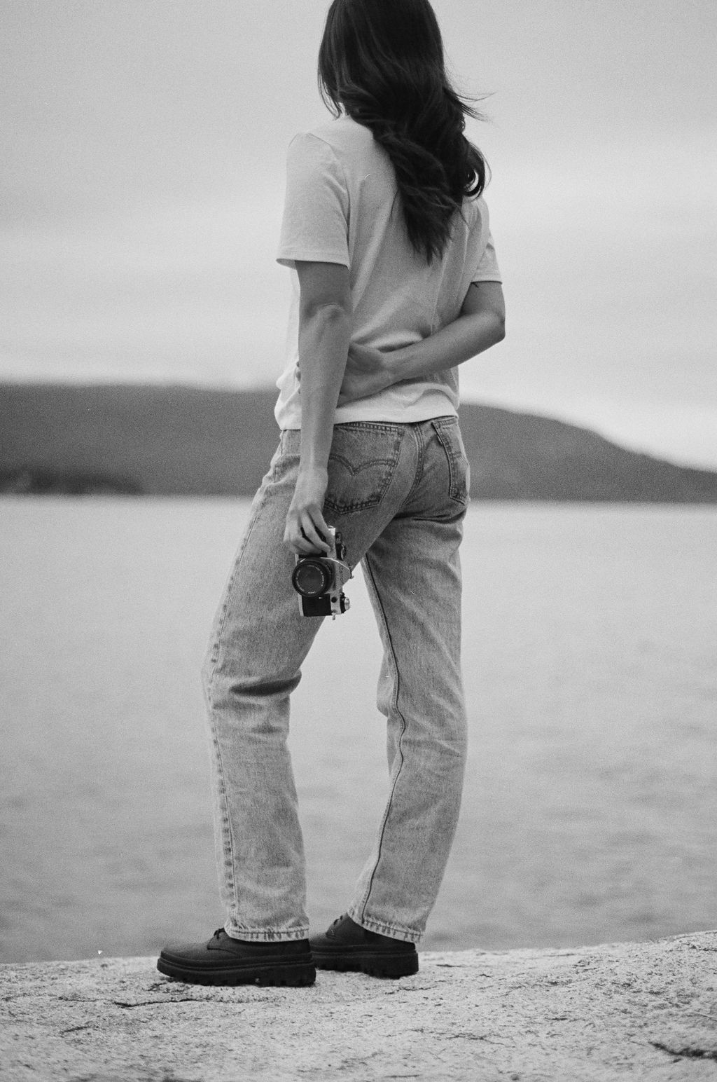 Woman walking barefoot on a sandy beach with shallow water, wearing jeans and white shirt, with trees in background.
