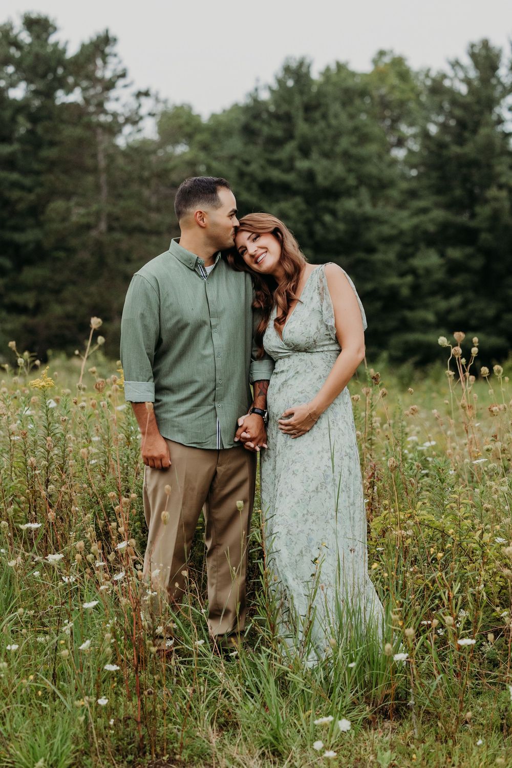 A couple stands in a wildflower meadow, the man kissing her head as she smiles, cradling her baby bump in a sage dress.