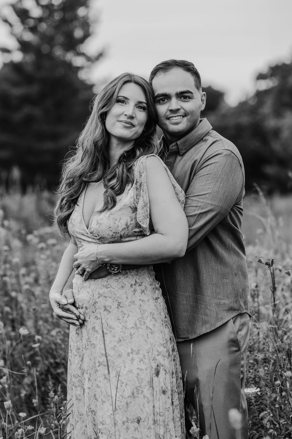 Black and white photo of a smiling couple embracing in a wildflower field, woman in floral dress, man in button-up shirt.