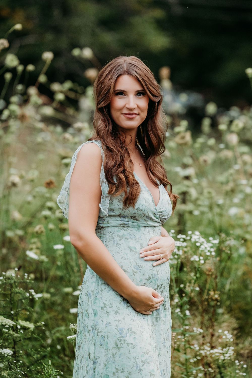 Pregnant woman in mint floral dress standing in wildflower field, cradling baby bump, with long auburn hair.
