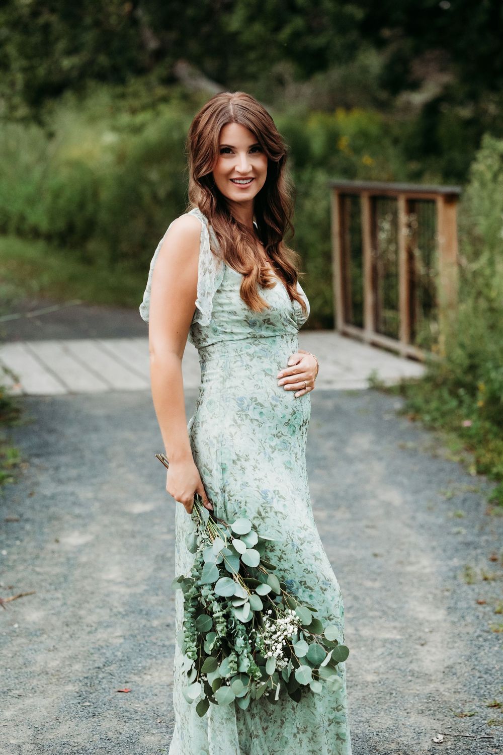 Smiling pregnant woman in green floral maxi dress holding eucalyptus bouquet on garden path.
