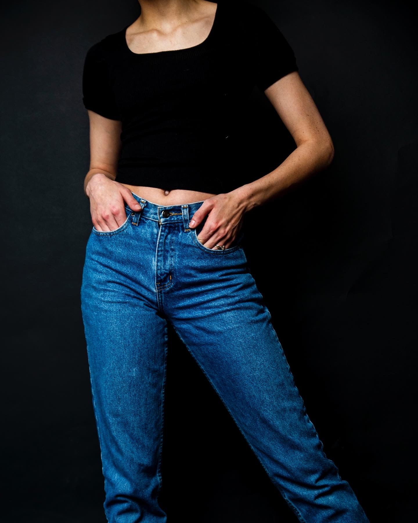 Woman wearing blue high-waist jeans and black crop top posing against dark background.
