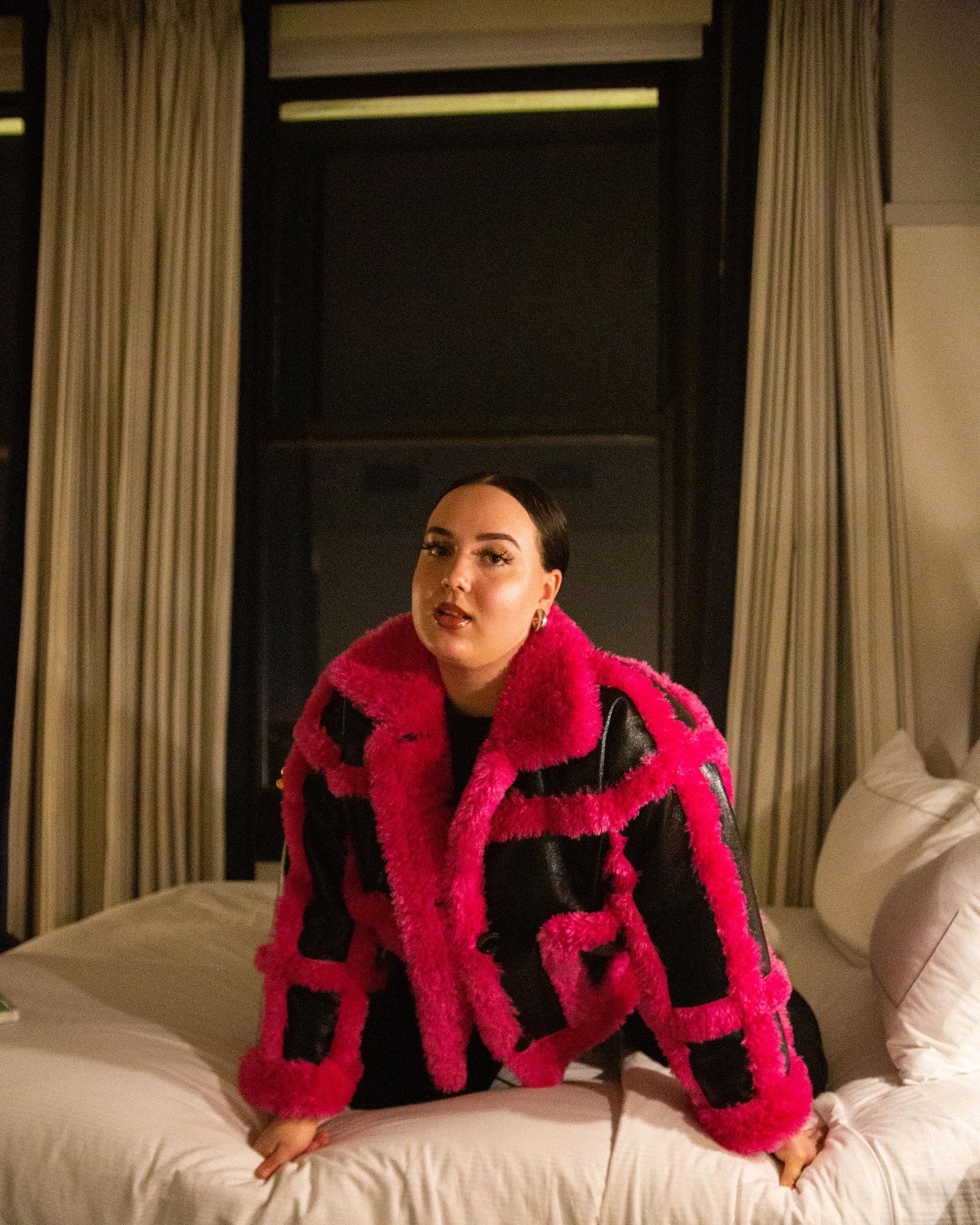 Woman posing on bed wearing bold pink and black faux fur coat in dimly lit hotel room.