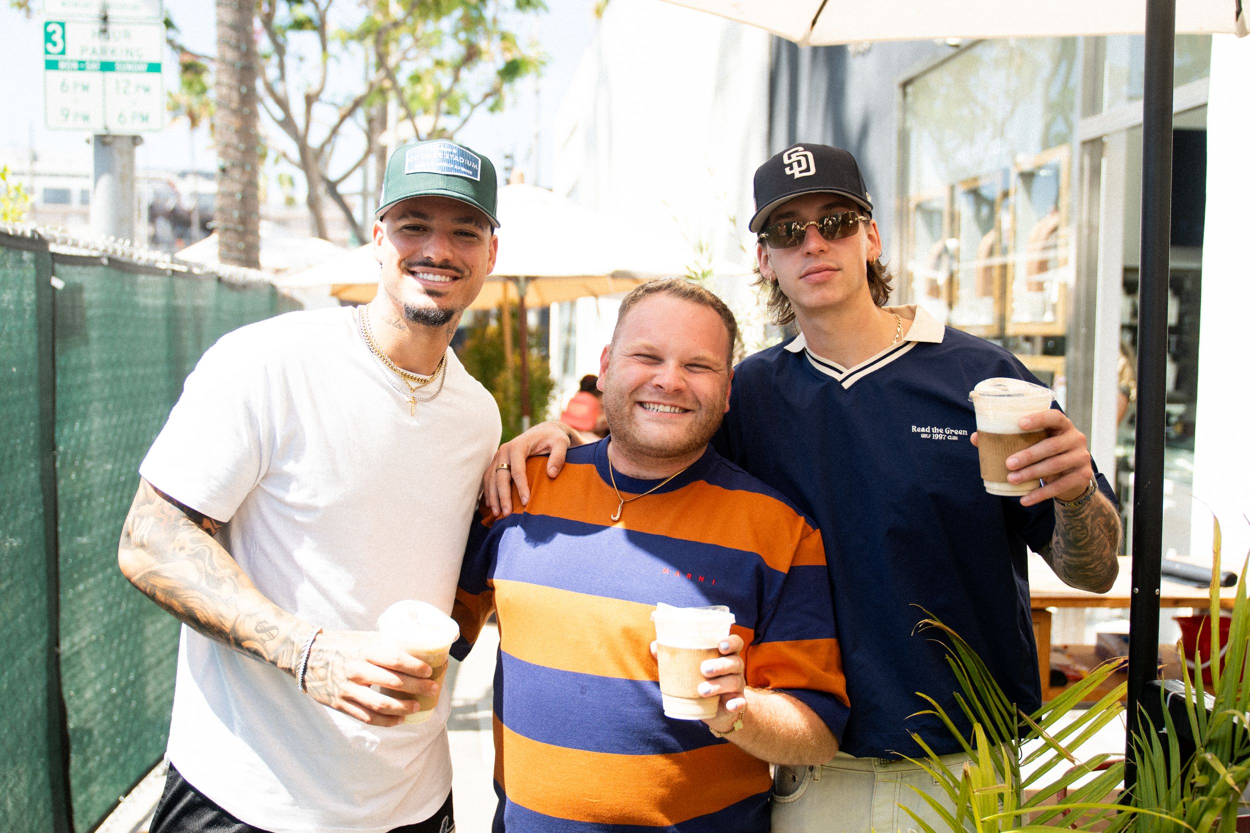 Three friends smiling and holding coffee cups outdoors at a sunny cafe patio, two with tattoos wearing caps.