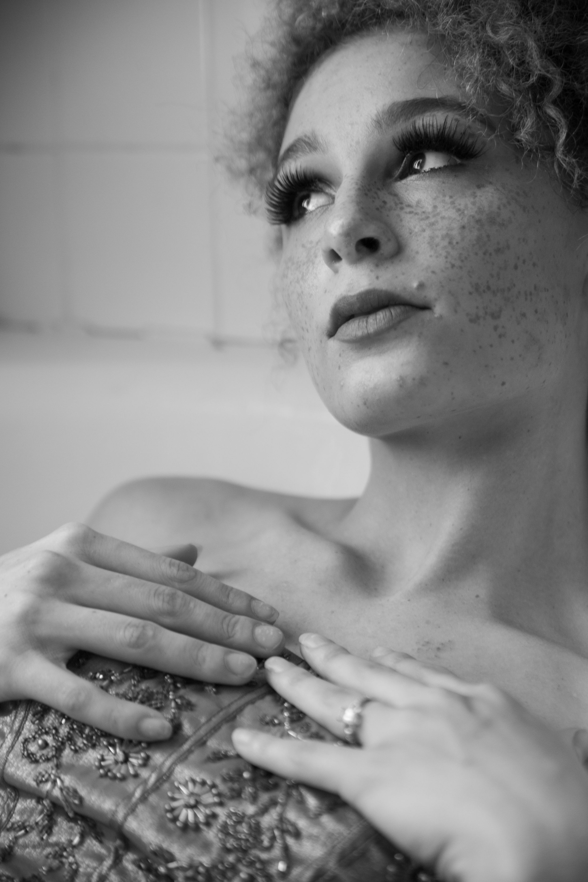 Black and white portrait of a young freckled woman with curly hair and dramatic lashes, hands crossed over chest.