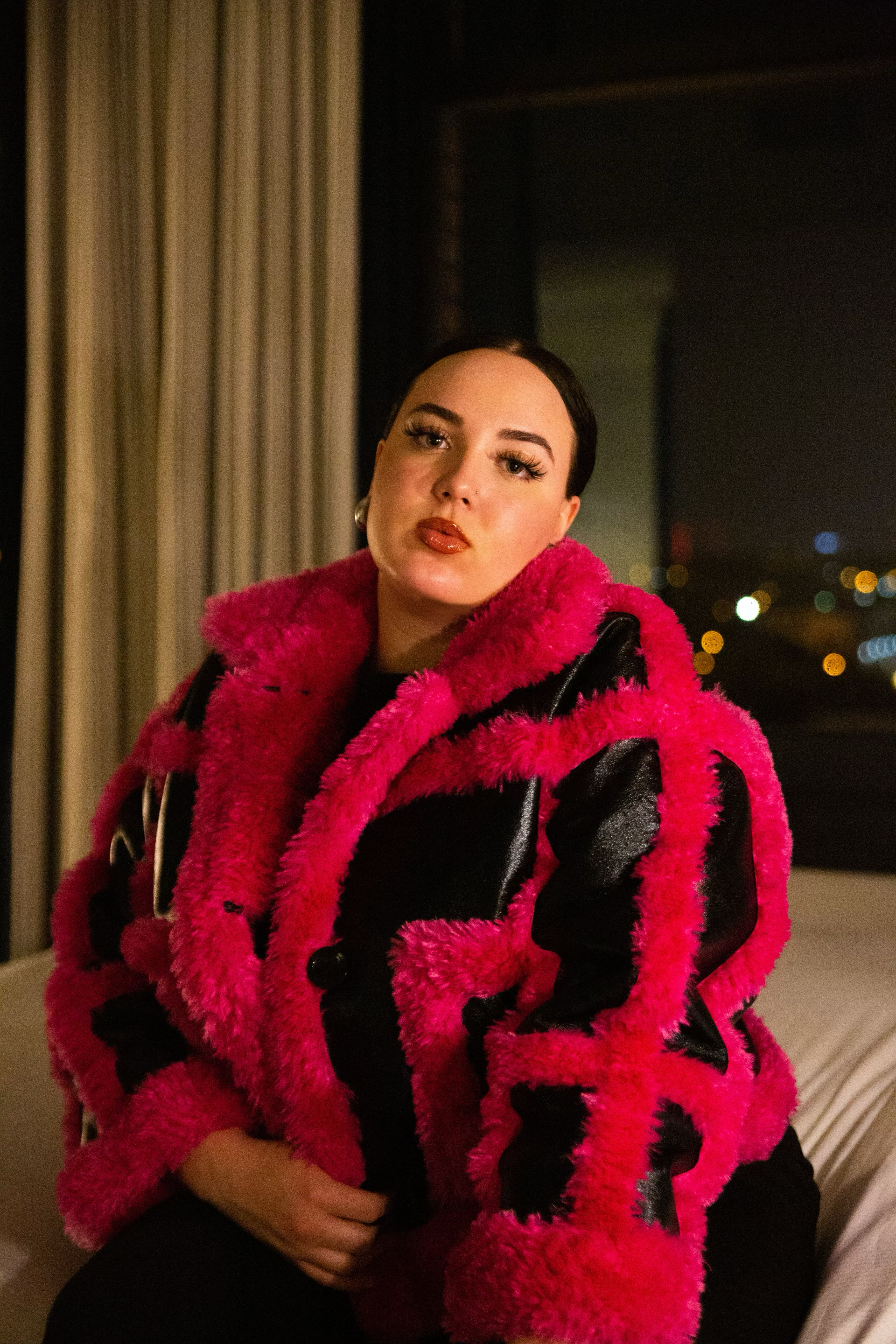 Woman in bold pink and black faux fur coat poses indoors with city lights glowing in background at night.
