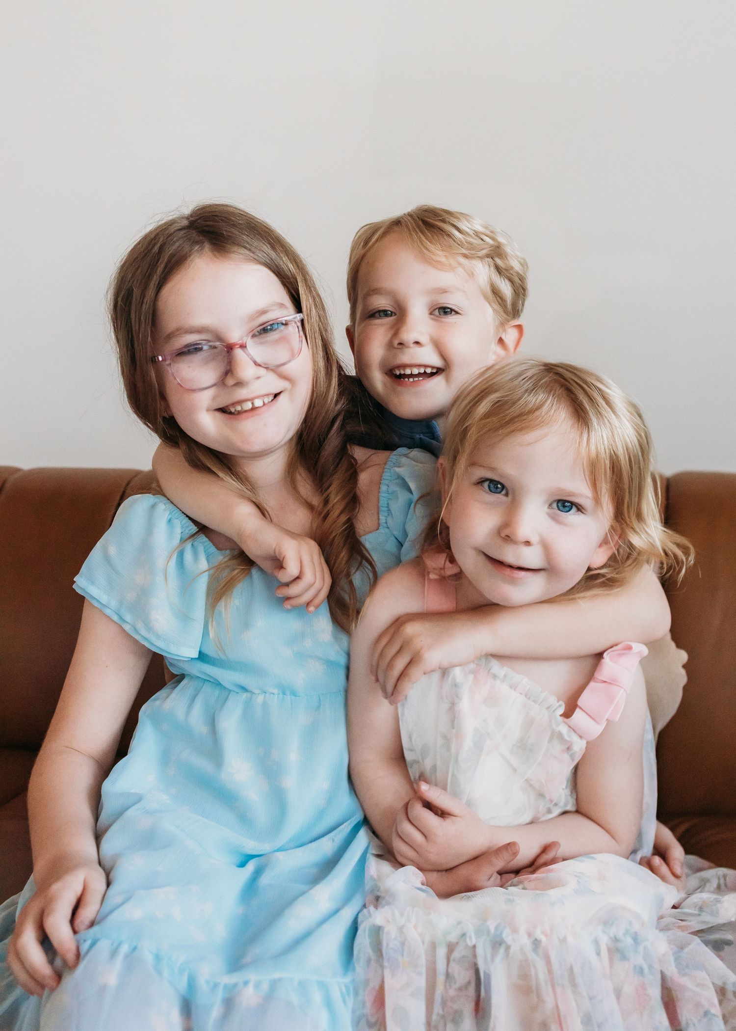 Three smiling children in pastel dresses pose together on a brown leather couch for a portrait photo.