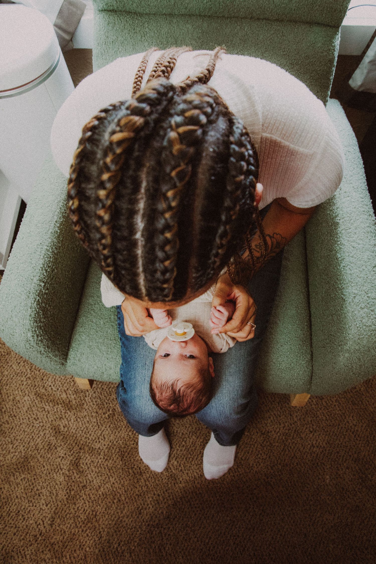 Overhead view of braided-hair mother cradling newborn baby in mint green armchair.