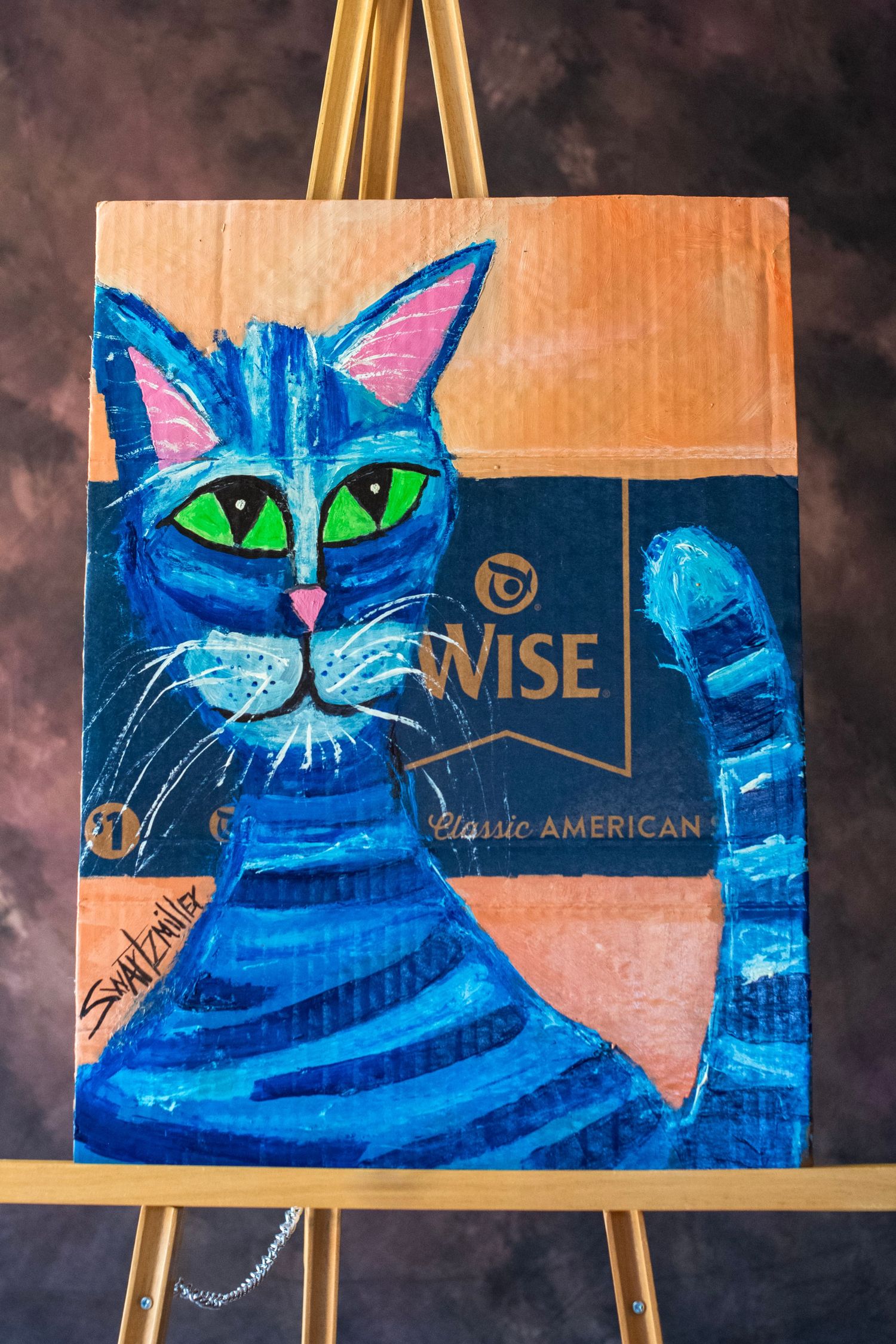 Blue cat painting on Wise chips cardboard, vibrant folk art with green eyes on an easel.