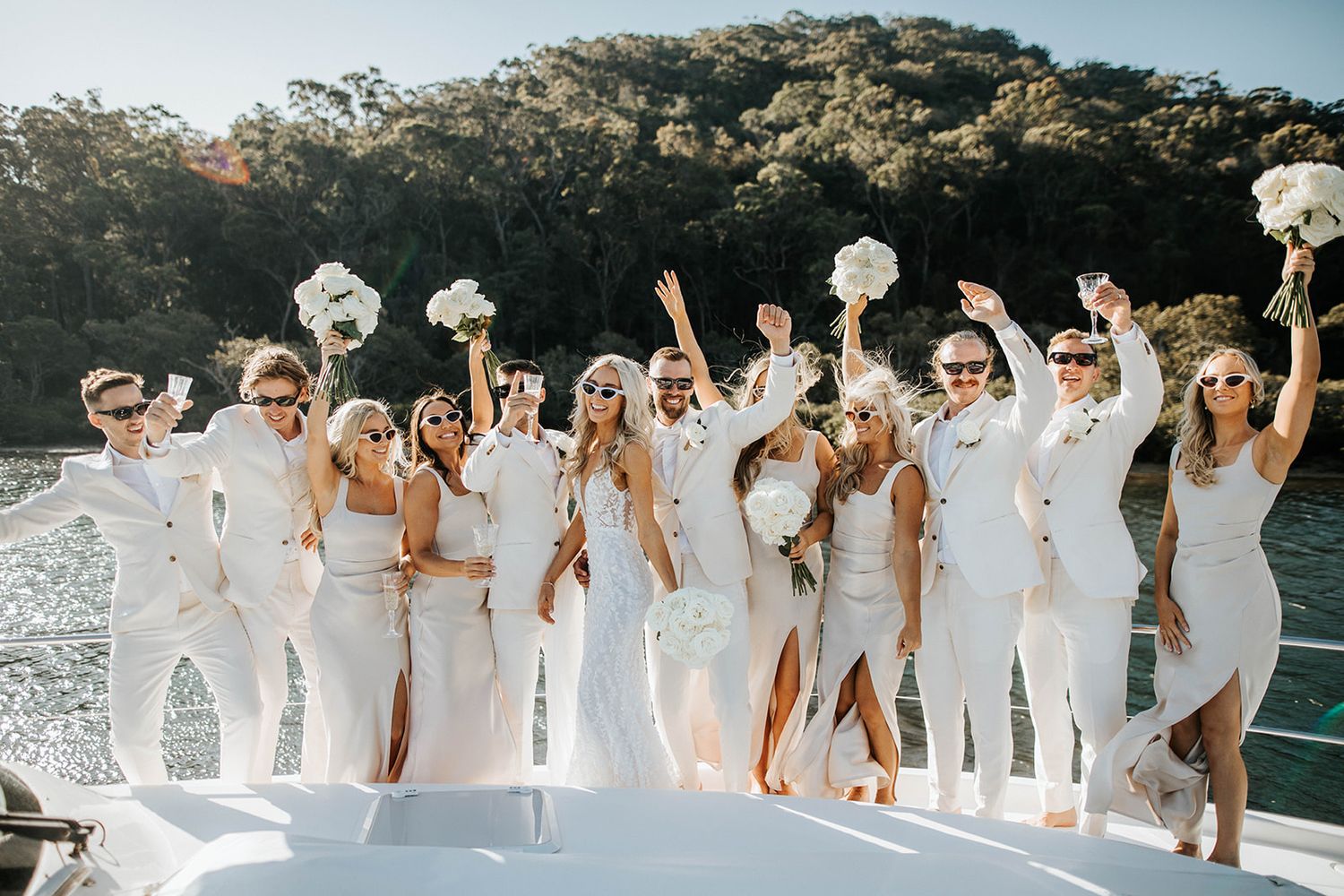 Wedding party in white attire celebrating on a boat deck with bouquets raised, lush green trees and water in background.