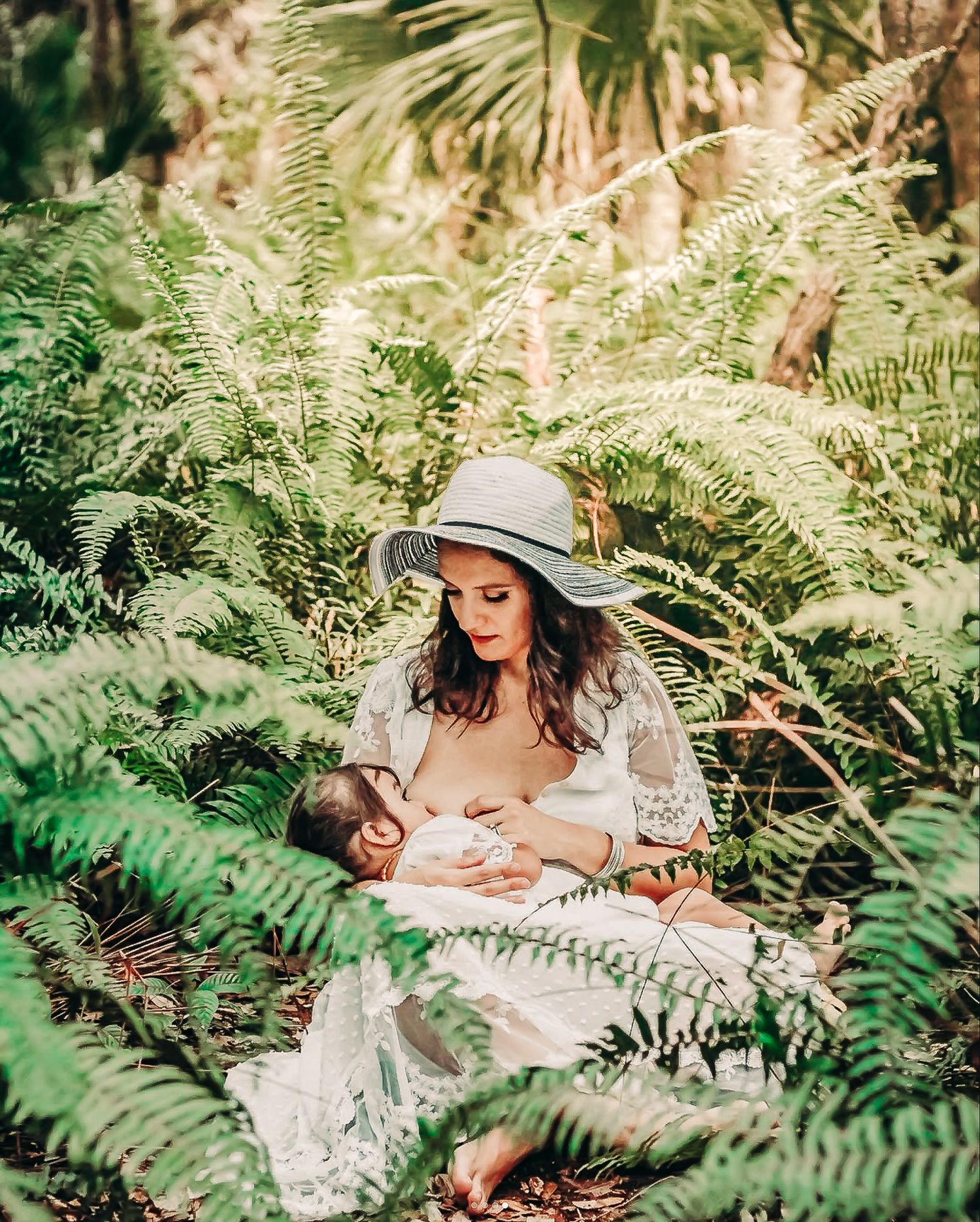A mother in a white lace dress and sun hat breastfeeds her newborn surrounded by lush green ferns in nature at Delray Oaks Natural Area.