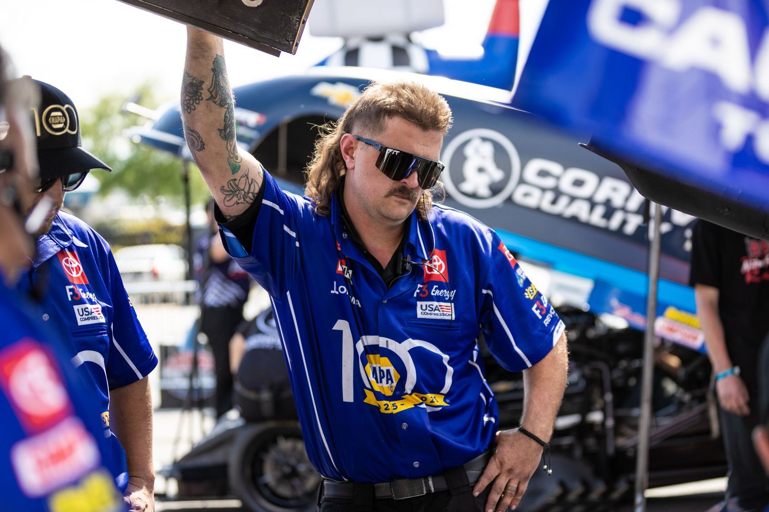 A tattooed NASCAR crew member in blue team uniform and sunglasses works at a racing event pit area.