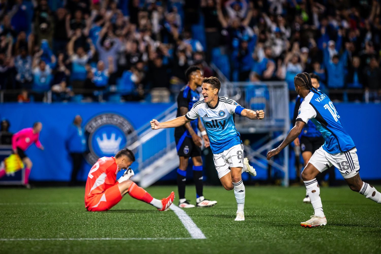 Soccer players celebrate a goal during an MLS match, with a goalkeeper on the ground in the background.