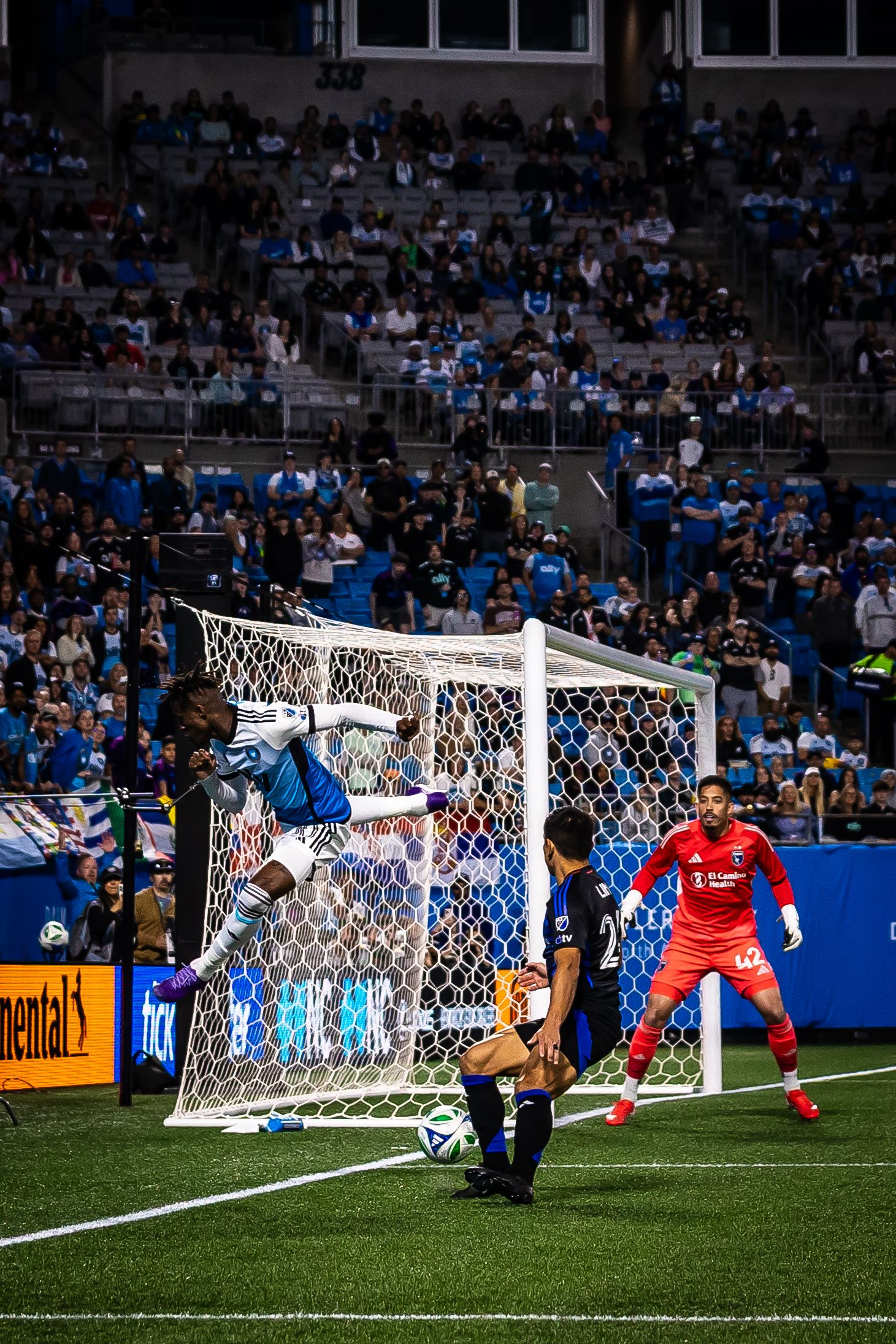 Soccer player leaping near goal net during MLS night match with goalkeeper and crowd in background.