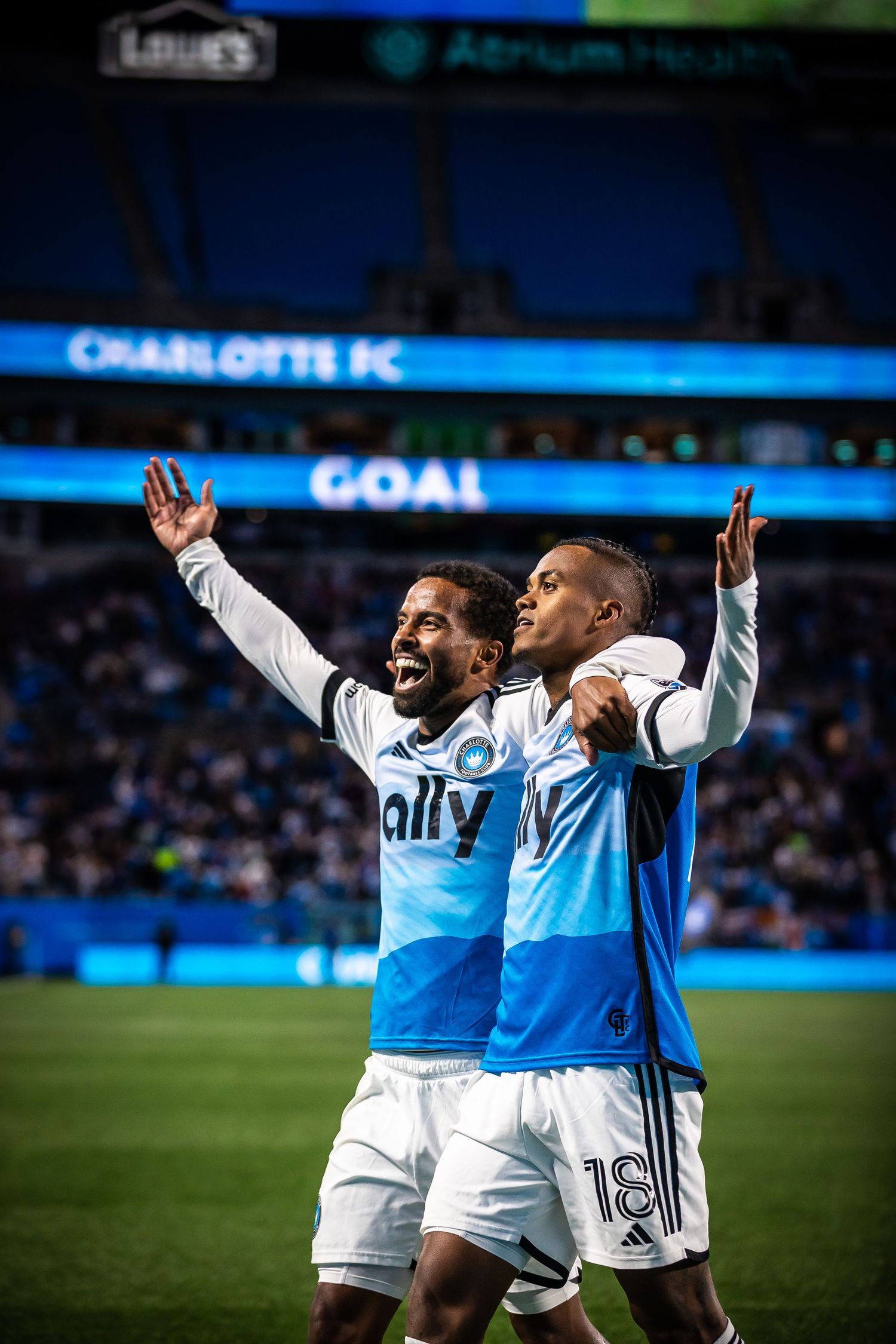 Charlotte FC players celebrate a goal on the field, wearing blue and white jerseys, with GOAL displayed on the stadium scoreboard.