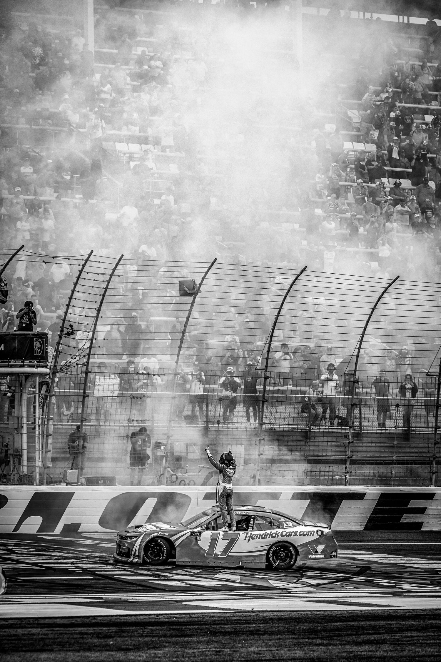 Black and white NASCAR race car burnout celebration with driver standing on roof amid smoke, packed grandstands visible behind fence.