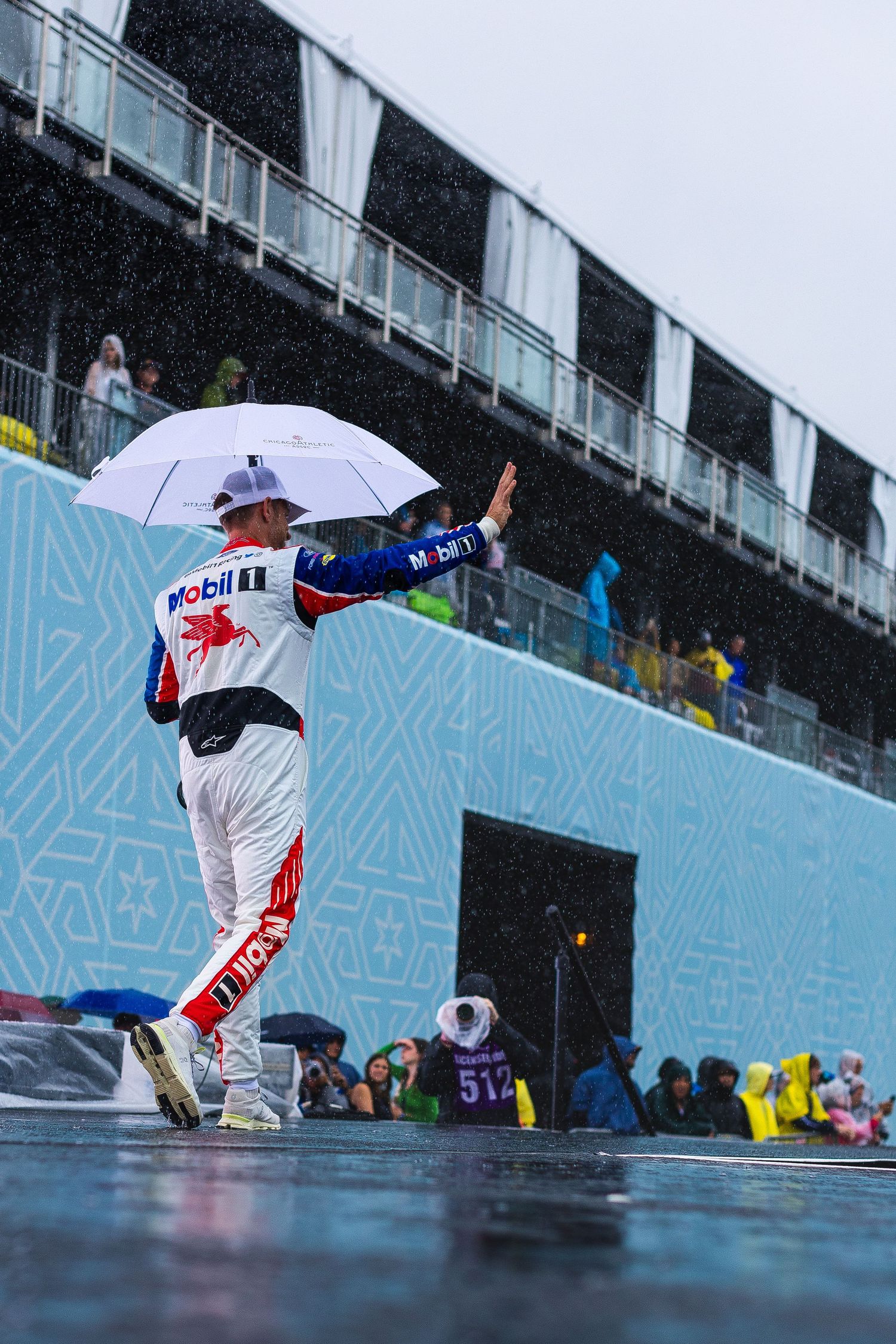 A racing driver in Mobil 1 livery walks in heavy rain holding a white umbrella at a motorsport event.