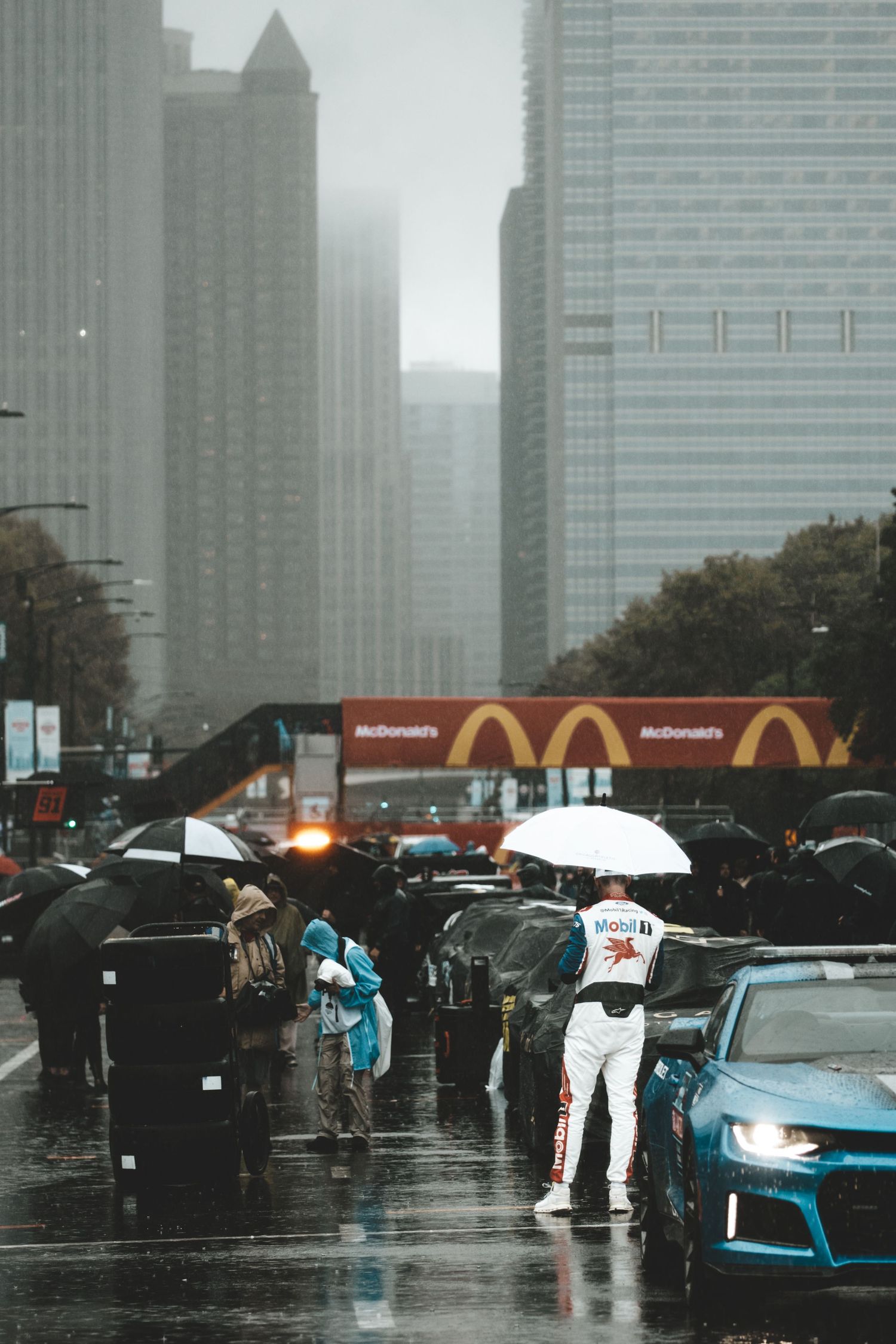 Racer in white suit holds umbrella beside blue sports car on rainy city street with McDonald's sign in background.