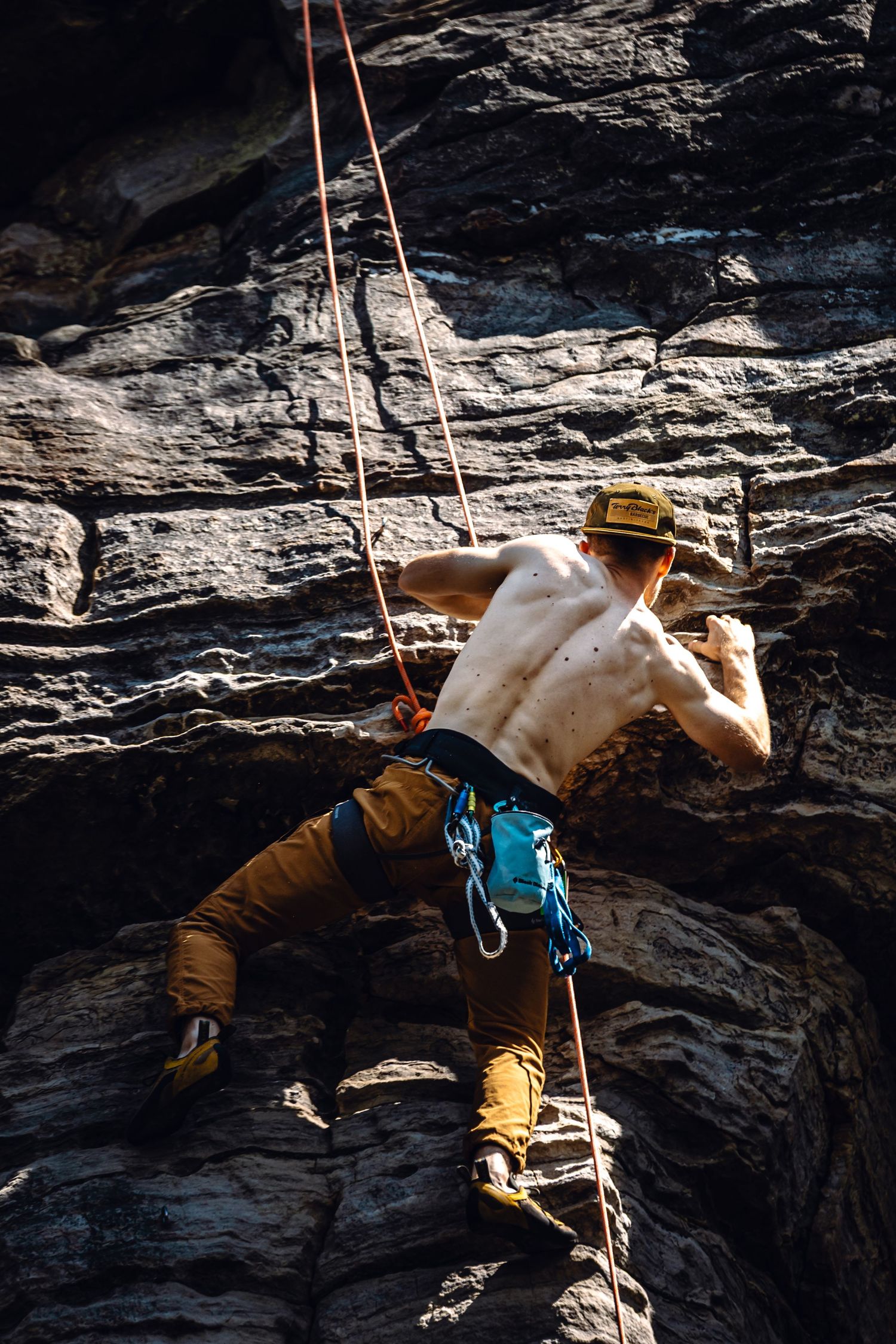 Shirtless male rock climber with safety harness and rope scaling a rugged dark stone cliff face outdoors.