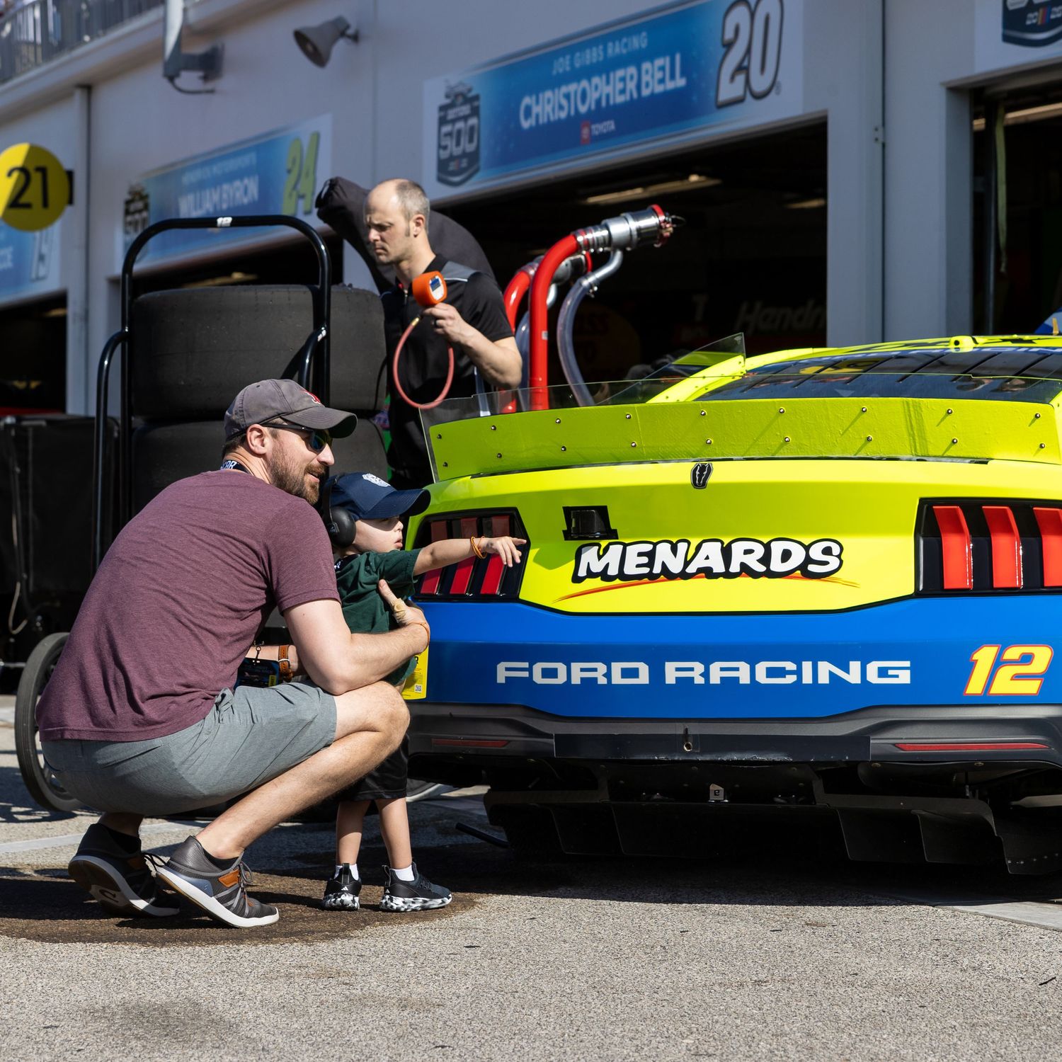 Man and child examine a blue and green Ford Racing Menards #12 NASCAR race car in the pit lane.