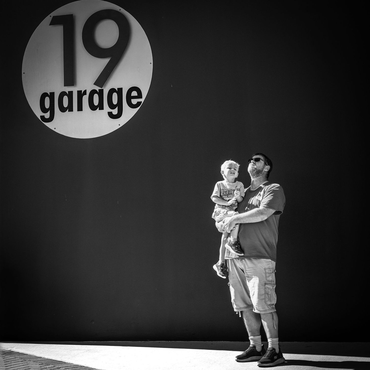 Man holding a child in front of a black wall with a circular sign reading 19 Garage, in black and white.