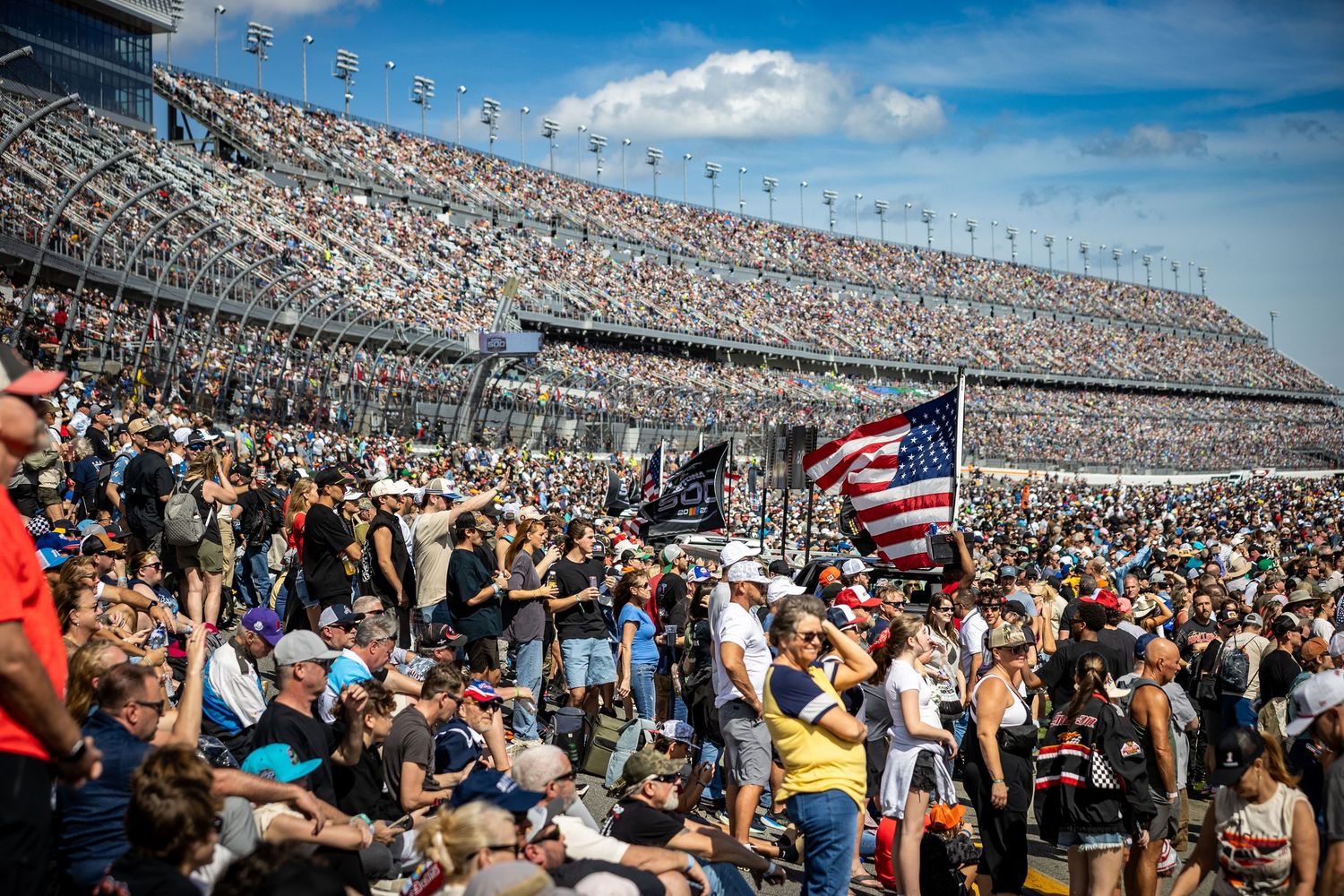 Massive crowd fills NASCAR Daytona 500 grandstands with American flag waving in foreground at packed racing event.