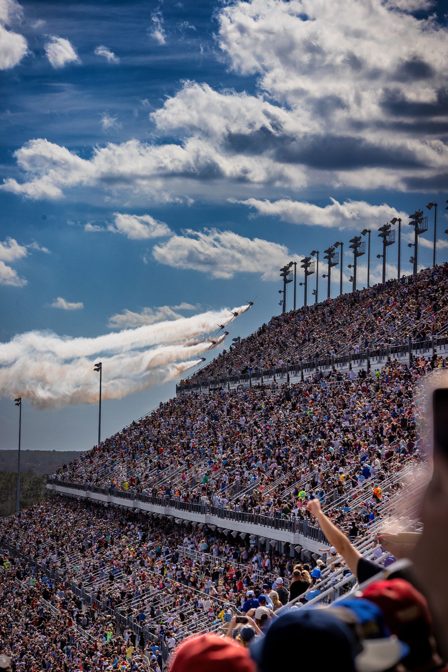 Military jets fly in formation over a packed NASCAR stadium grandstand under dramatic blue skies with white clouds.