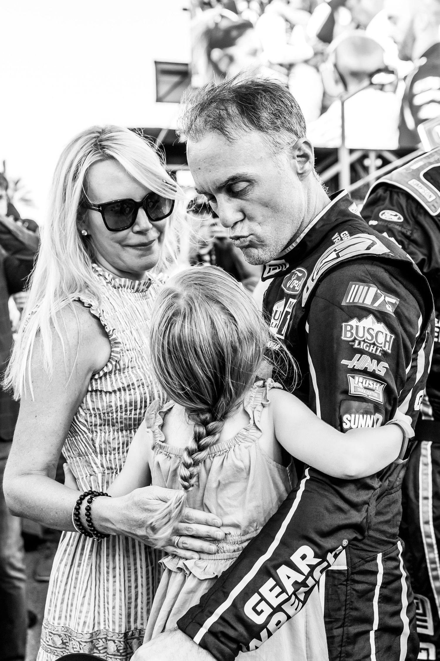 Black and white photo of a NASCAR driver in racing suit embracing his wife and young children at a race event.