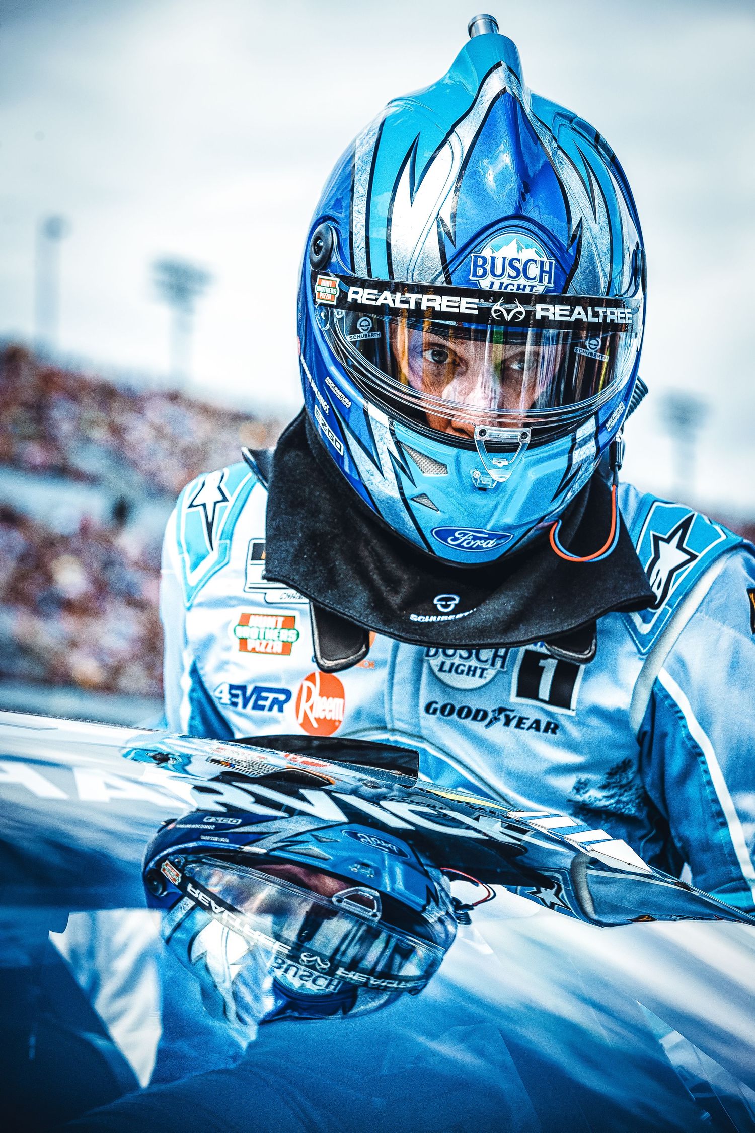 NASCAR driver in blue Busch Light Realtree helmet and racing suit leaning on car before race.