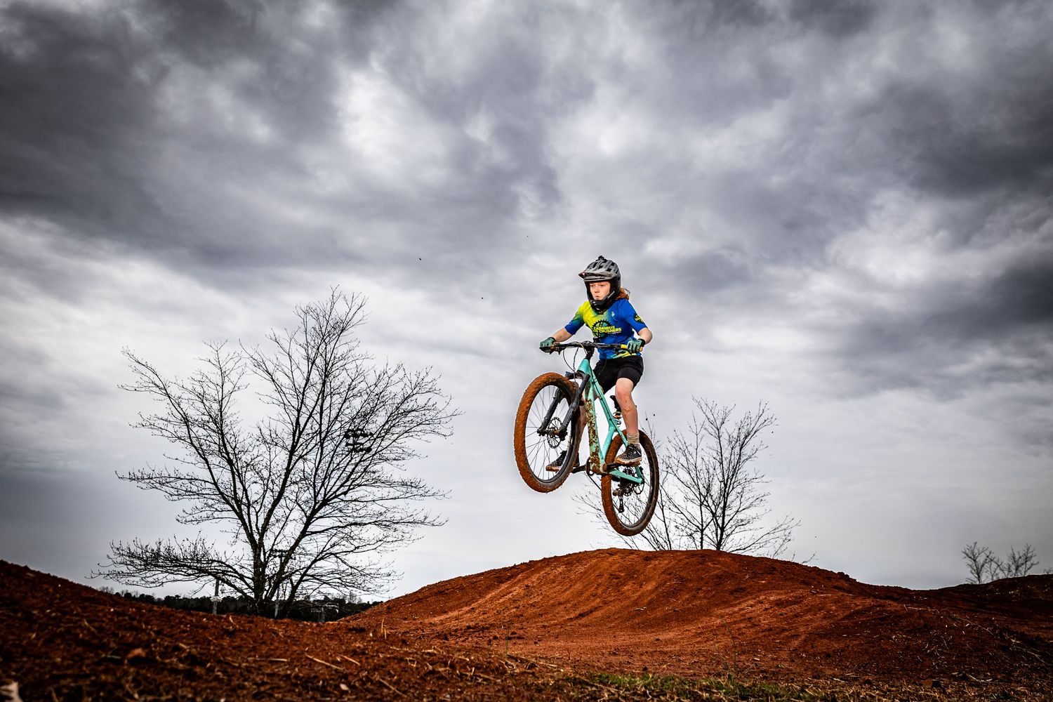 A young mountain biker catches air off a red dirt jump under dramatic cloudy skies with bare trees in the background.