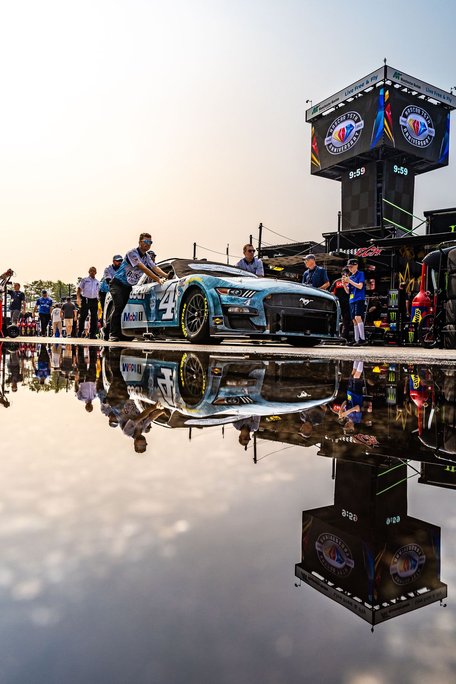 NASCAR Ford Mustang race car reflected in puddle on pit road during golden hour at a speedway event.