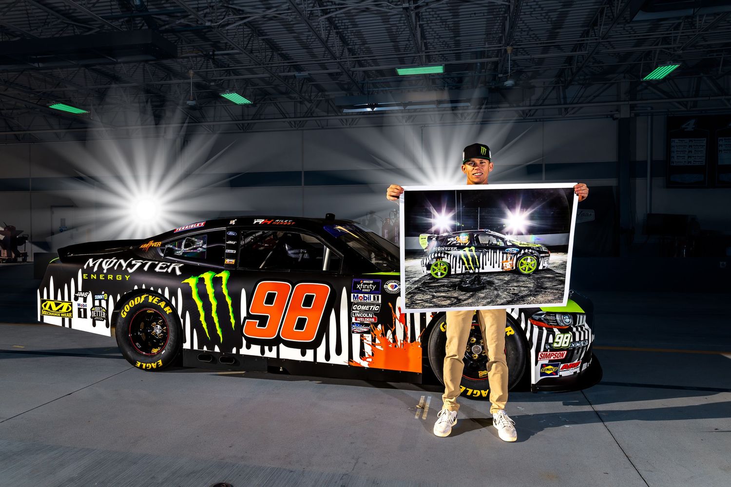 NASCAR driver holds a photo beside the Monster Energy #98 race car in a dark garage setting.