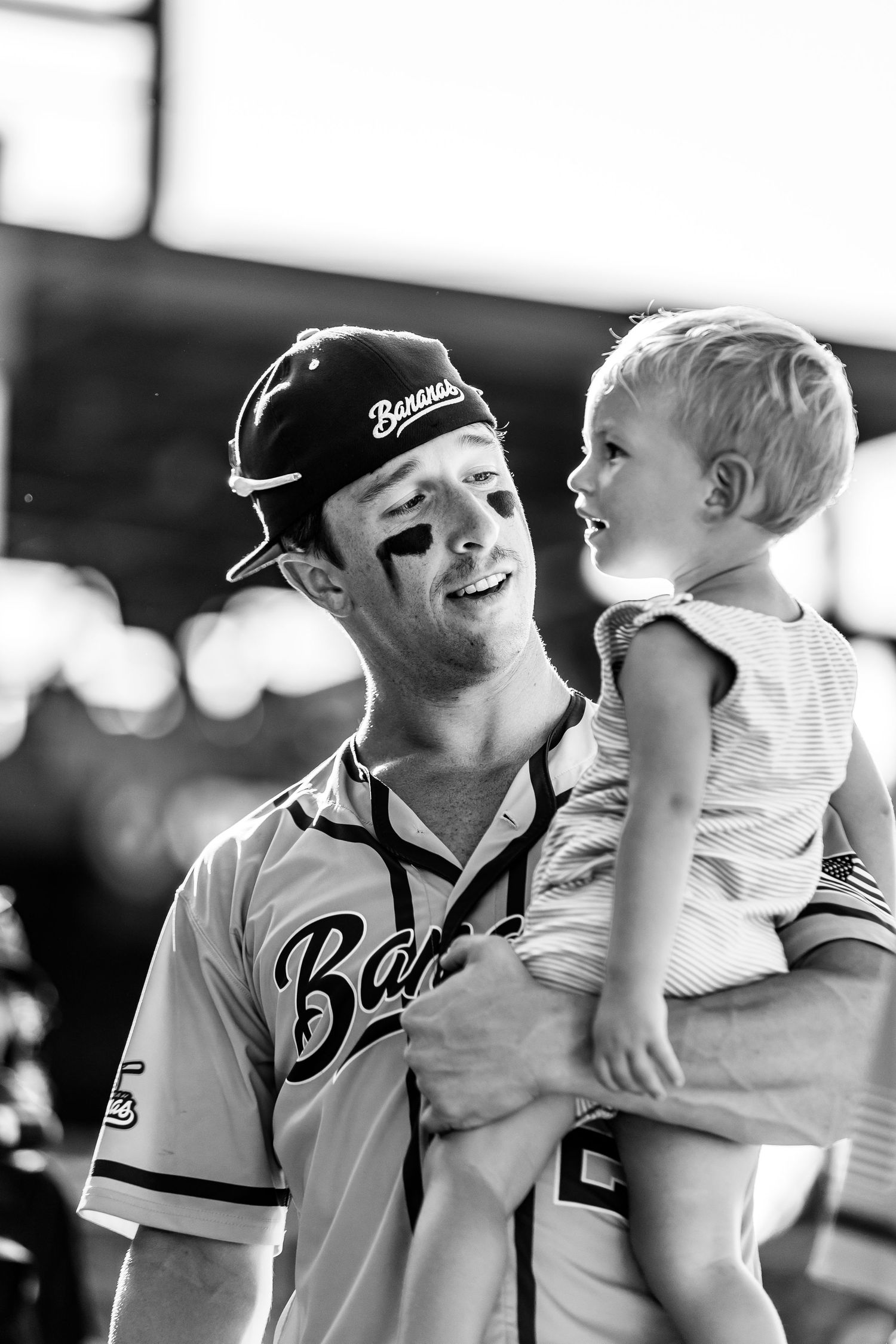 Baseball player in uniform holding a laughing toddler at a stadium in black and white.